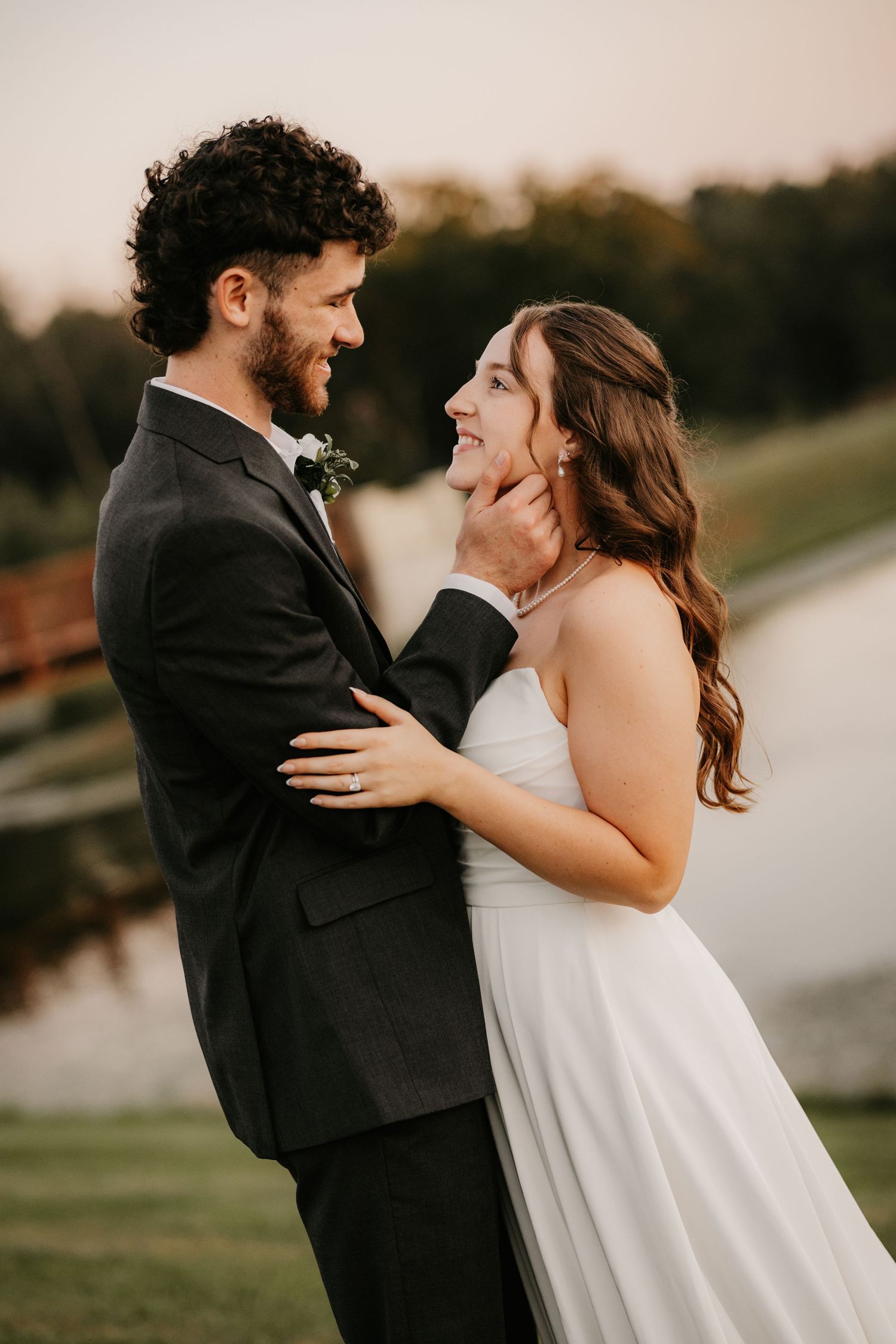 Bearded groom in a dark suit gently touches his bride's chin as the couple share a loving gaze beside a pond at sunset.