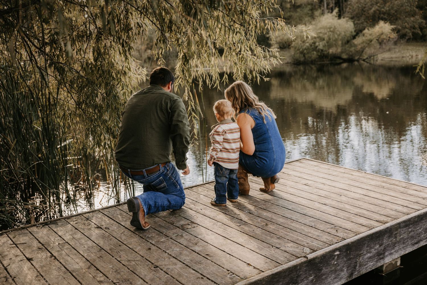 Father, mother, and toddler crouch together at the edge of a wooden dock, gazing at the lake surrounded by willow trees.