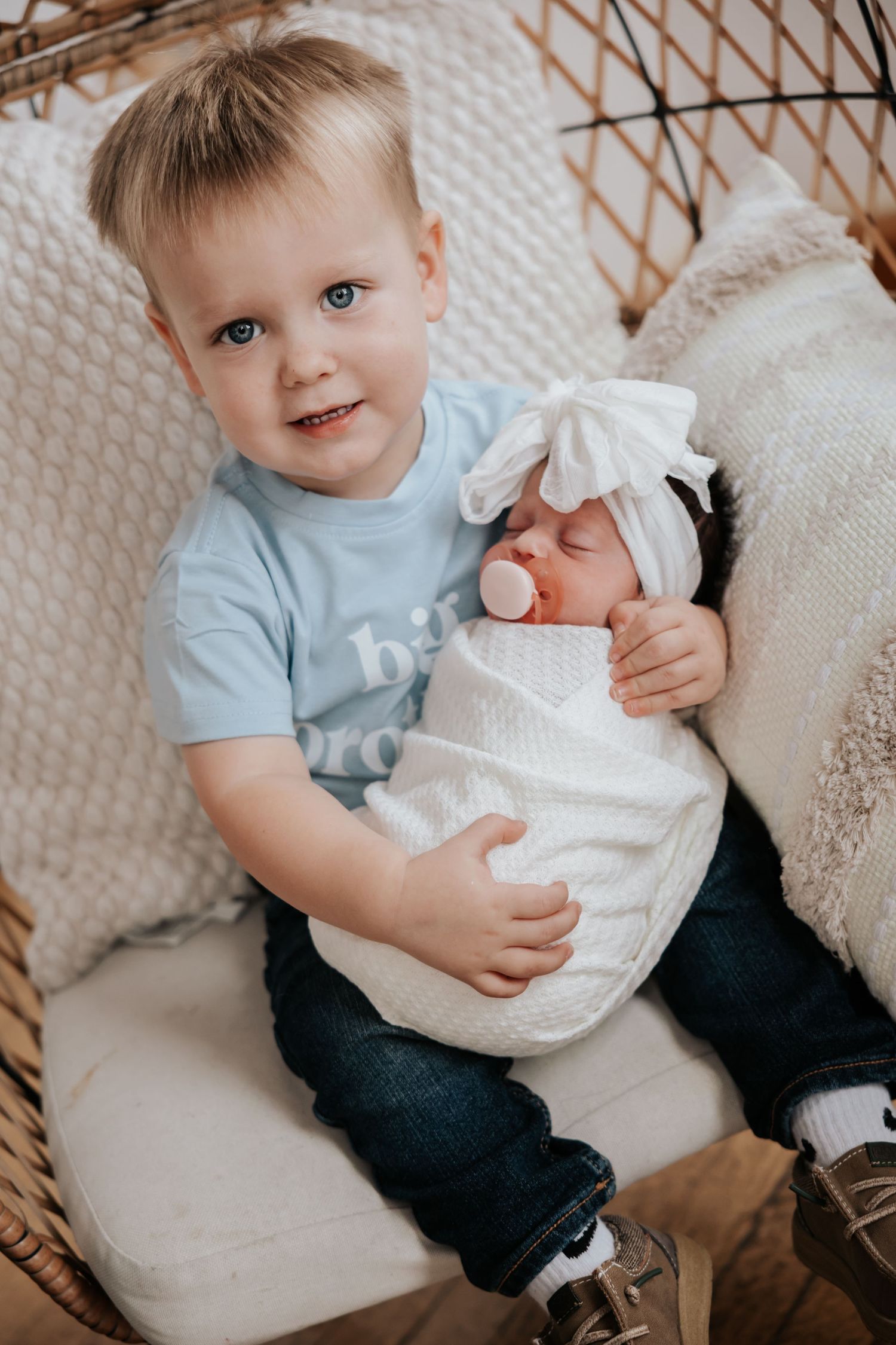 Smiling blond toddler in a big brother shirt holds his swaddled newborn sister with a pacifier and bow while seated in a wicker chair.