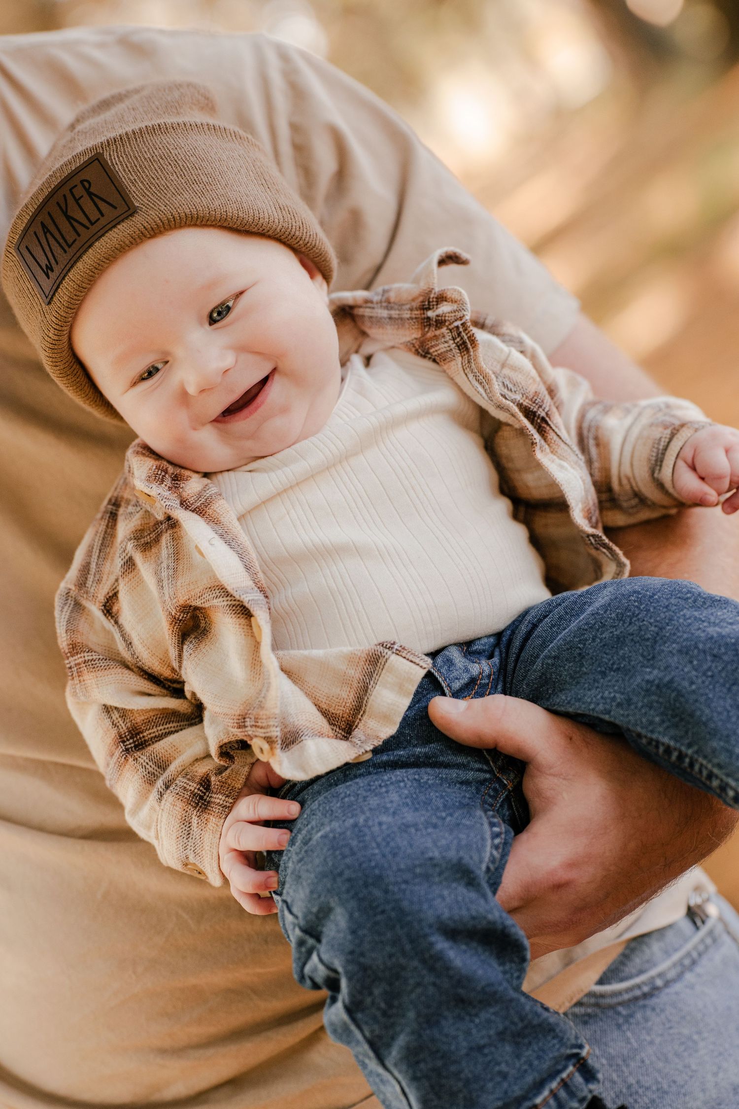 Smiling baby in a tan WALKER beanie and plaid flannel shirt is held up by a parent in a sunny autumn forest.