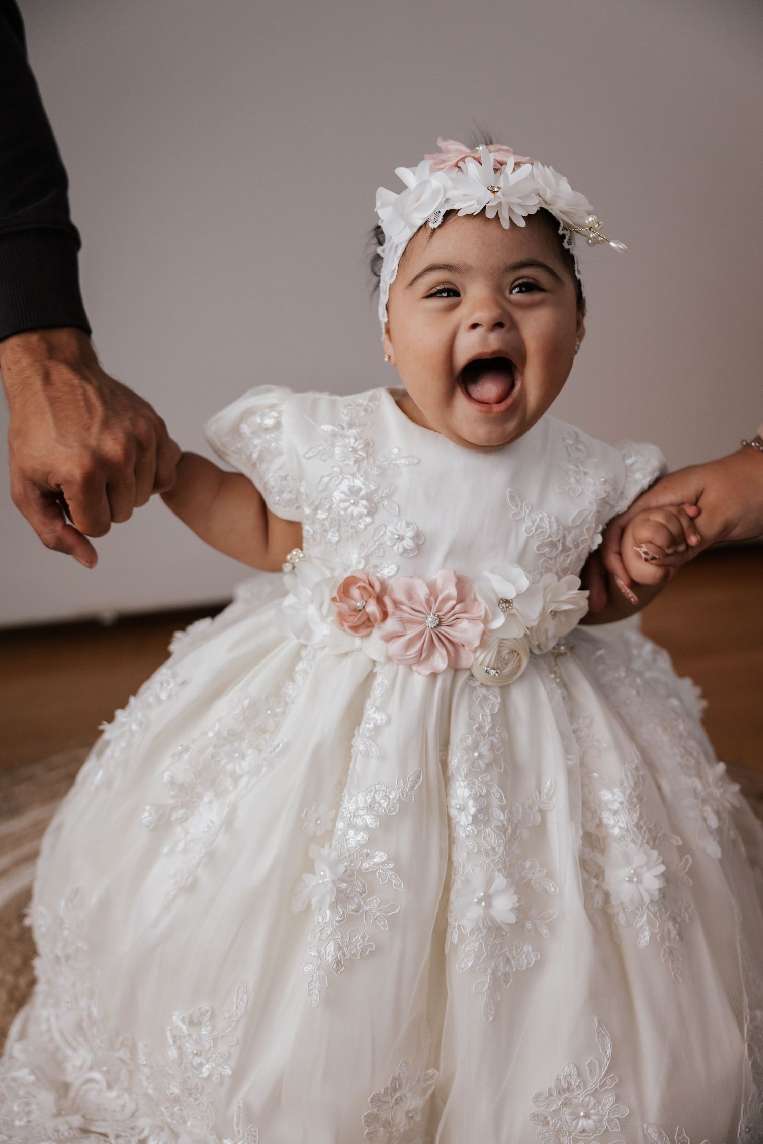 Excited baby in an ivory lace dress with pink flowers and a floral headband stands while two adults hold her hands.