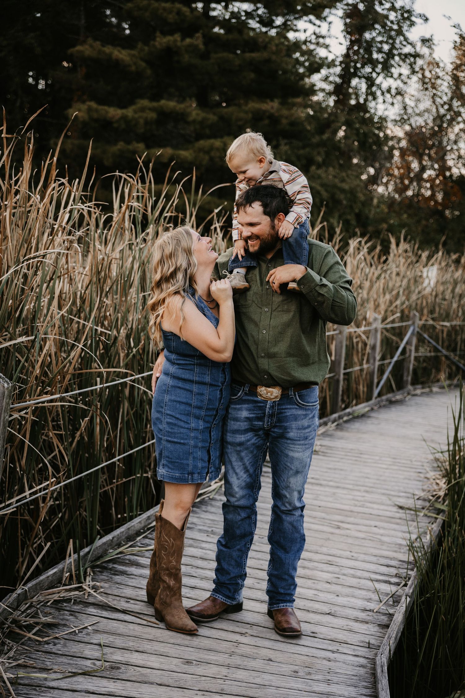 A laughing family stands on a wooden boardwalk by tall reeds as a dad carries his toddler on his shoulders.
