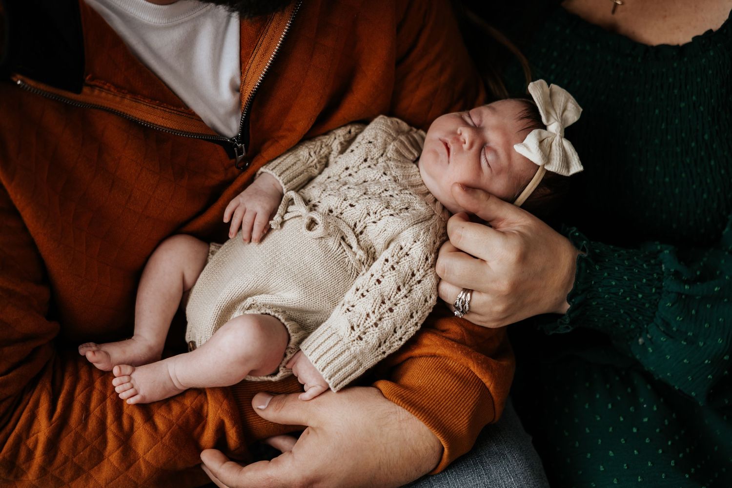 A close-up of a sleeping newborn in a cream knit romper and bow headband resting in her father's arms as her mother touches her.