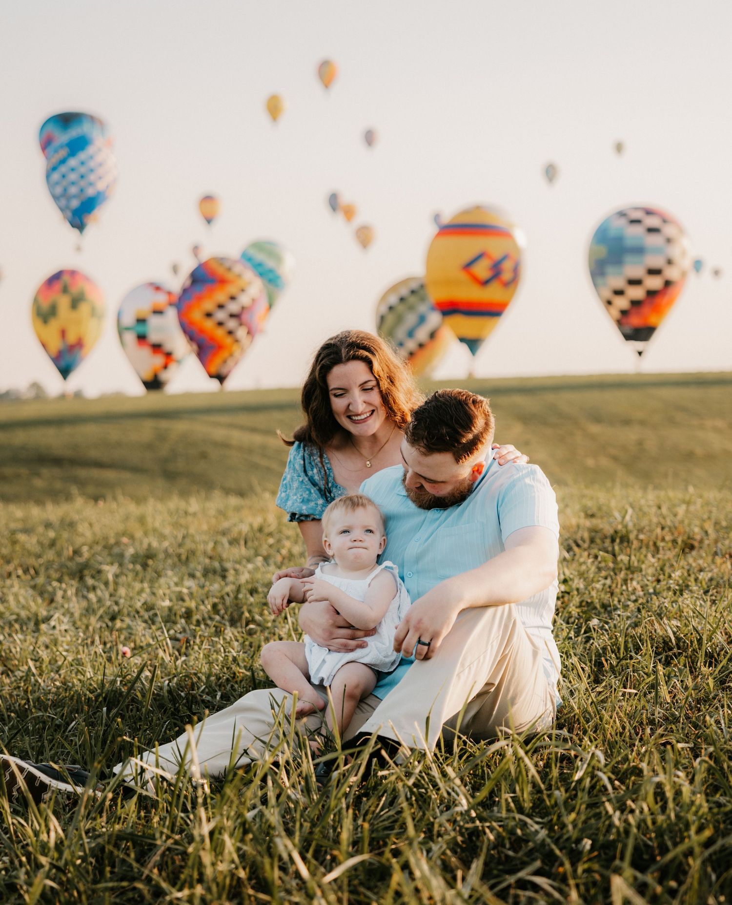 Smiling couple sits in a grassy field holding their baby while colorful hot air balloons float in the sky behind them.