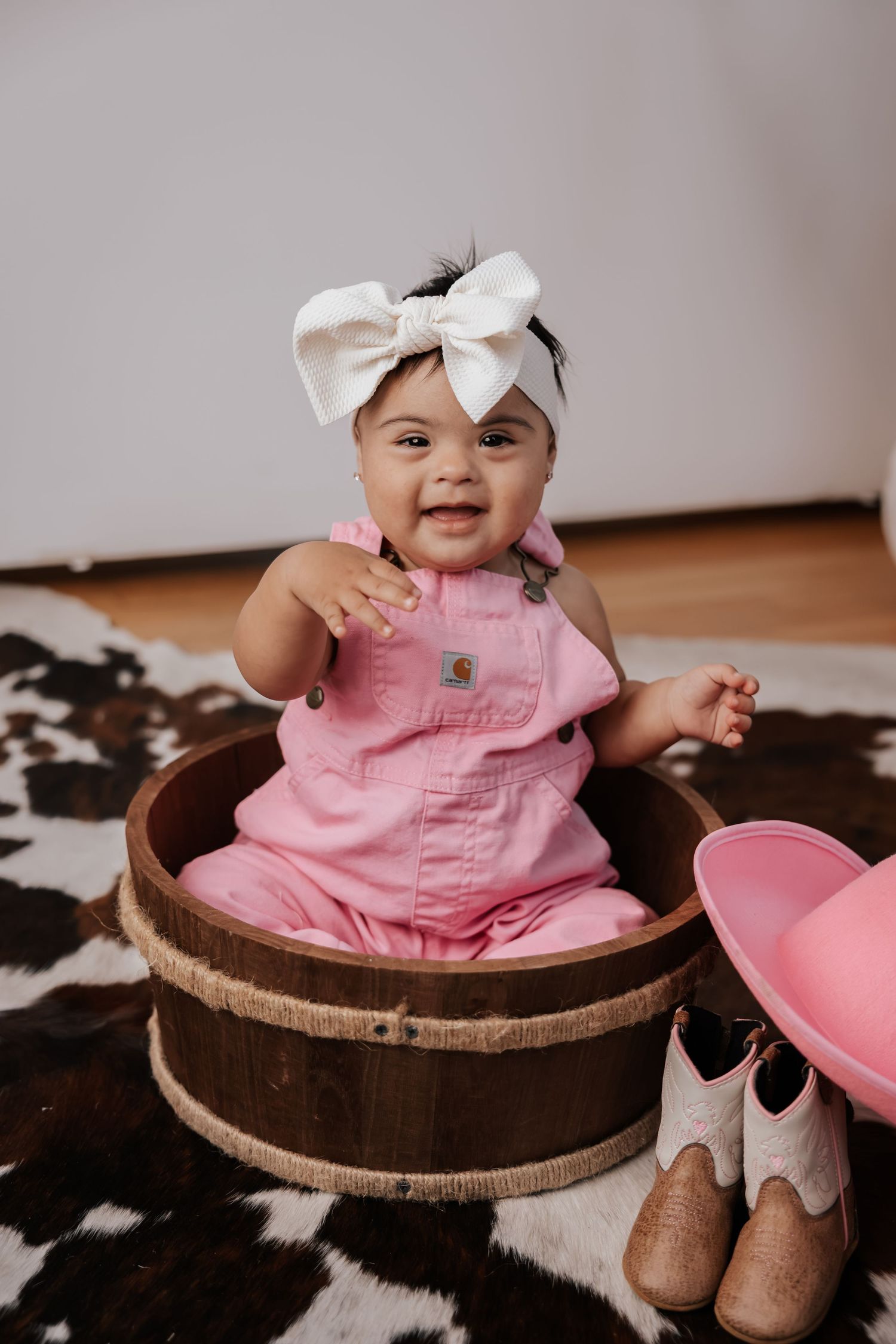 Smiling baby girl in pink overalls and a white bow headband sits in a wooden barrel beside cowgirl boots and a pink hat.