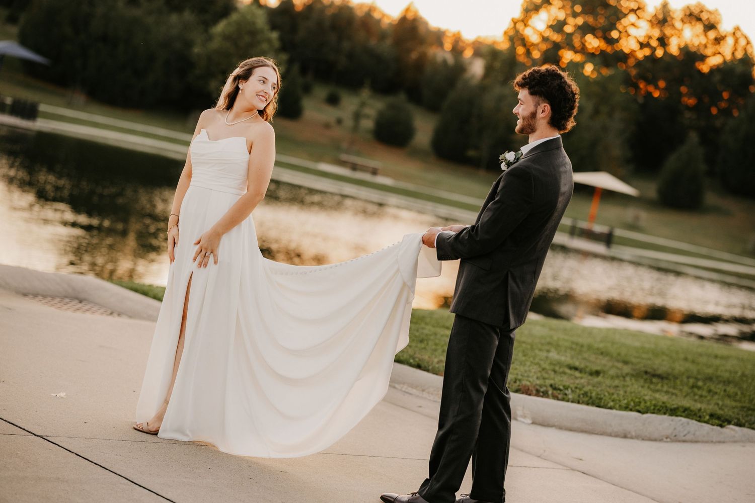 Bride in a flowing white gown and groom in a dark suit hold hands on a lakeside path at golden hour.