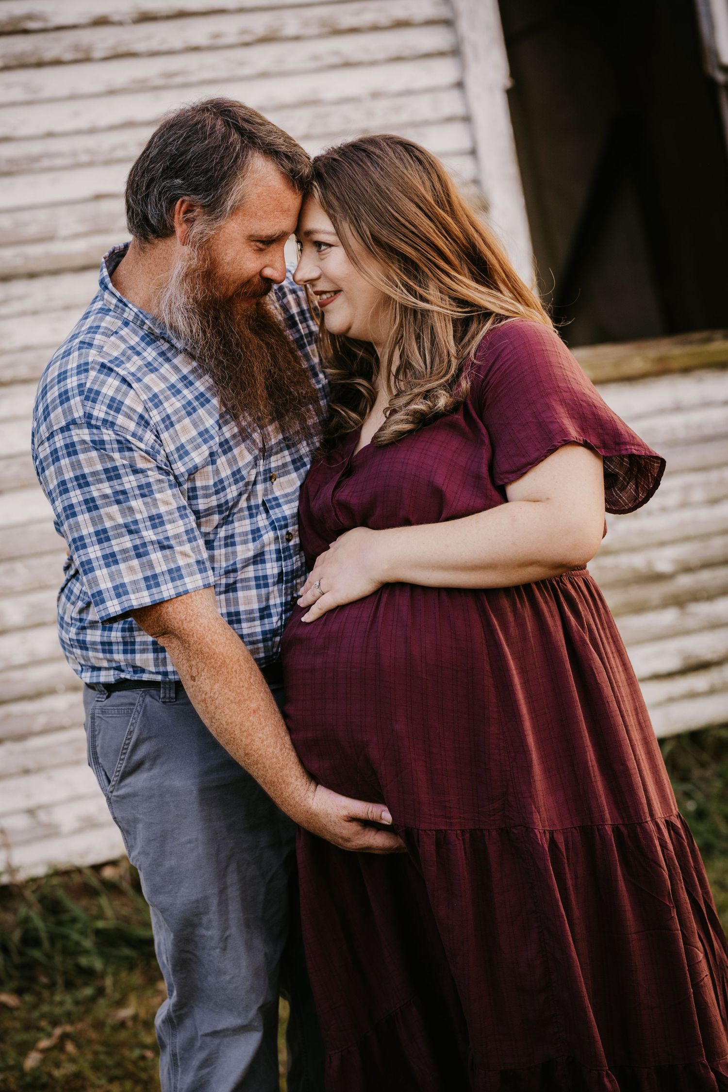 Bearded man in a blue plaid shirt gently touches his pregnant wife's belly as they share a tender moment by a rustic white barn.