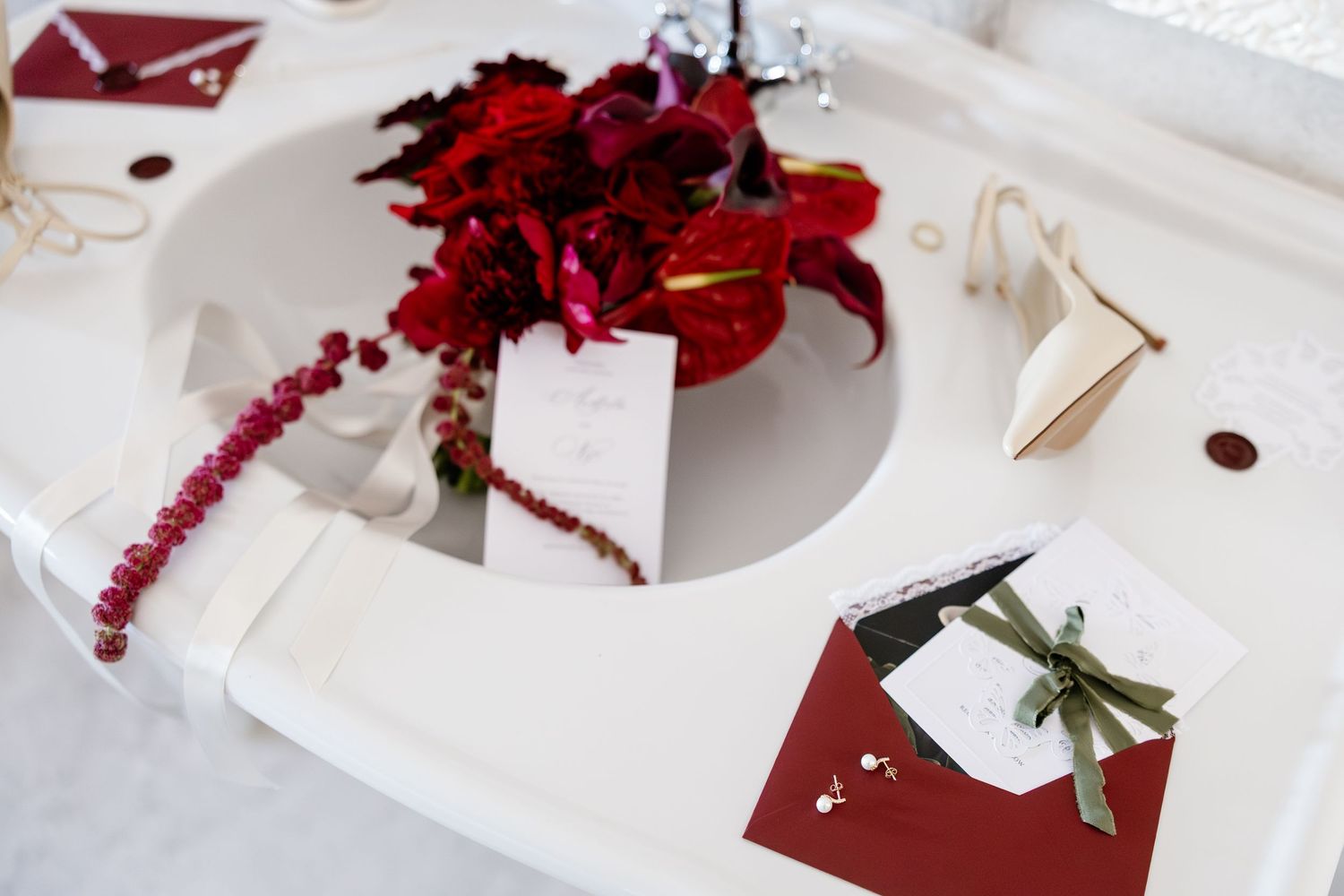Elegant wedding stationery with red floral arrangement, burgundy envelope, green ribbon, and pearl bracelet on white surface.