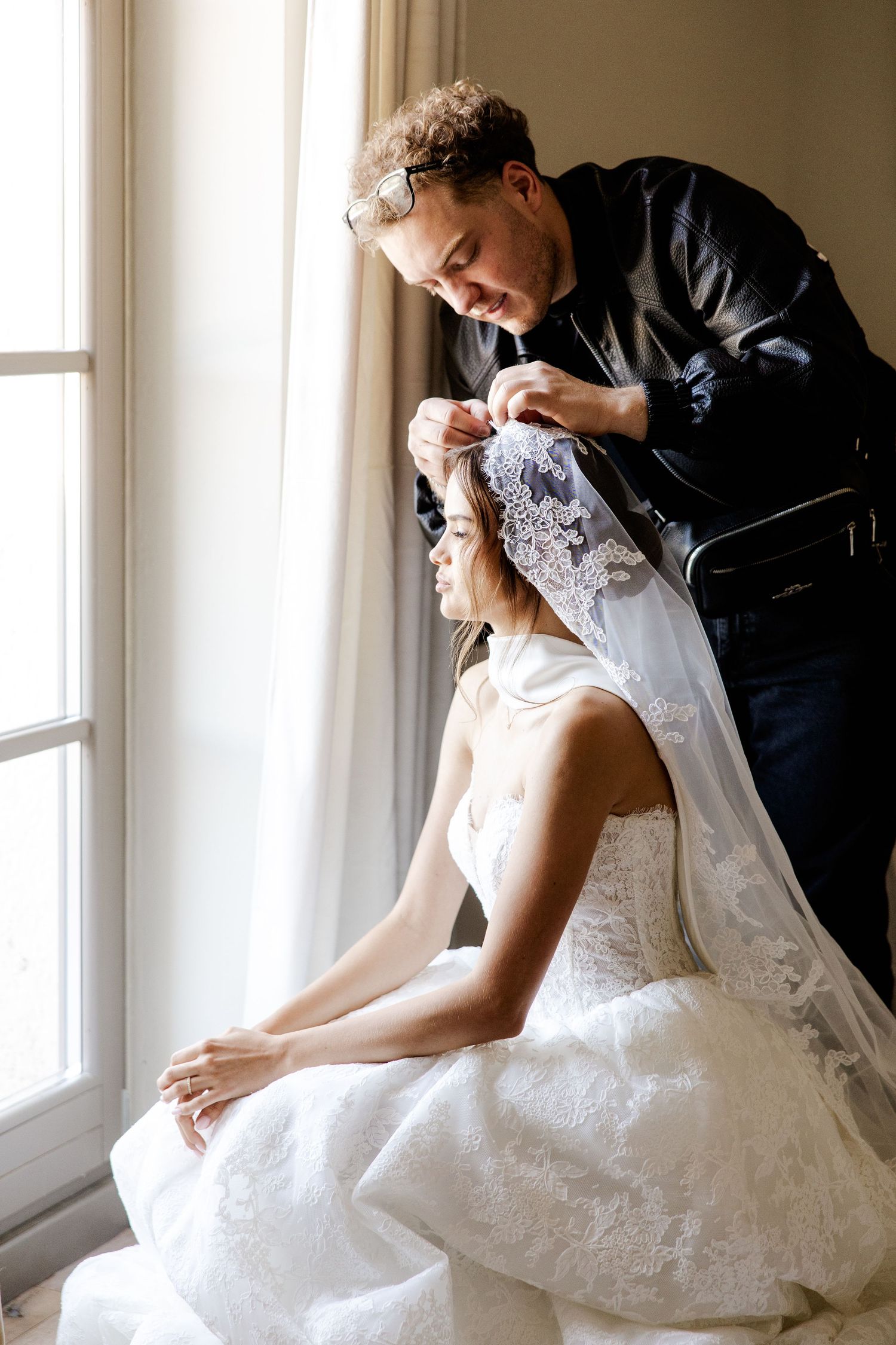 A hairstylist carefully attaches a lace bridal veil to a bride in a white wedding dress near a bright window.