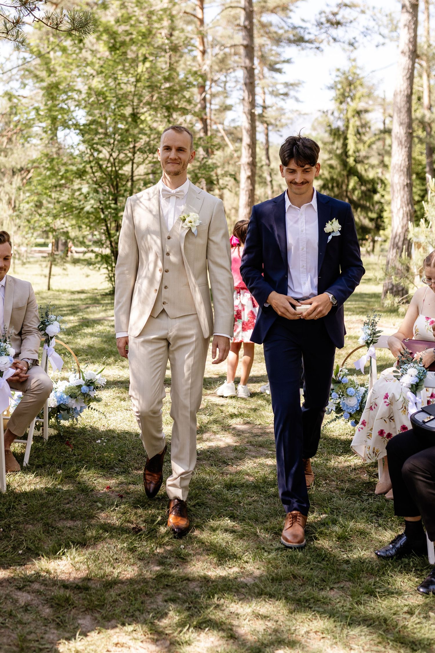 Groom in a beige suit walks down the outdoor wedding aisle accompanied by a groomsman in navy blue.