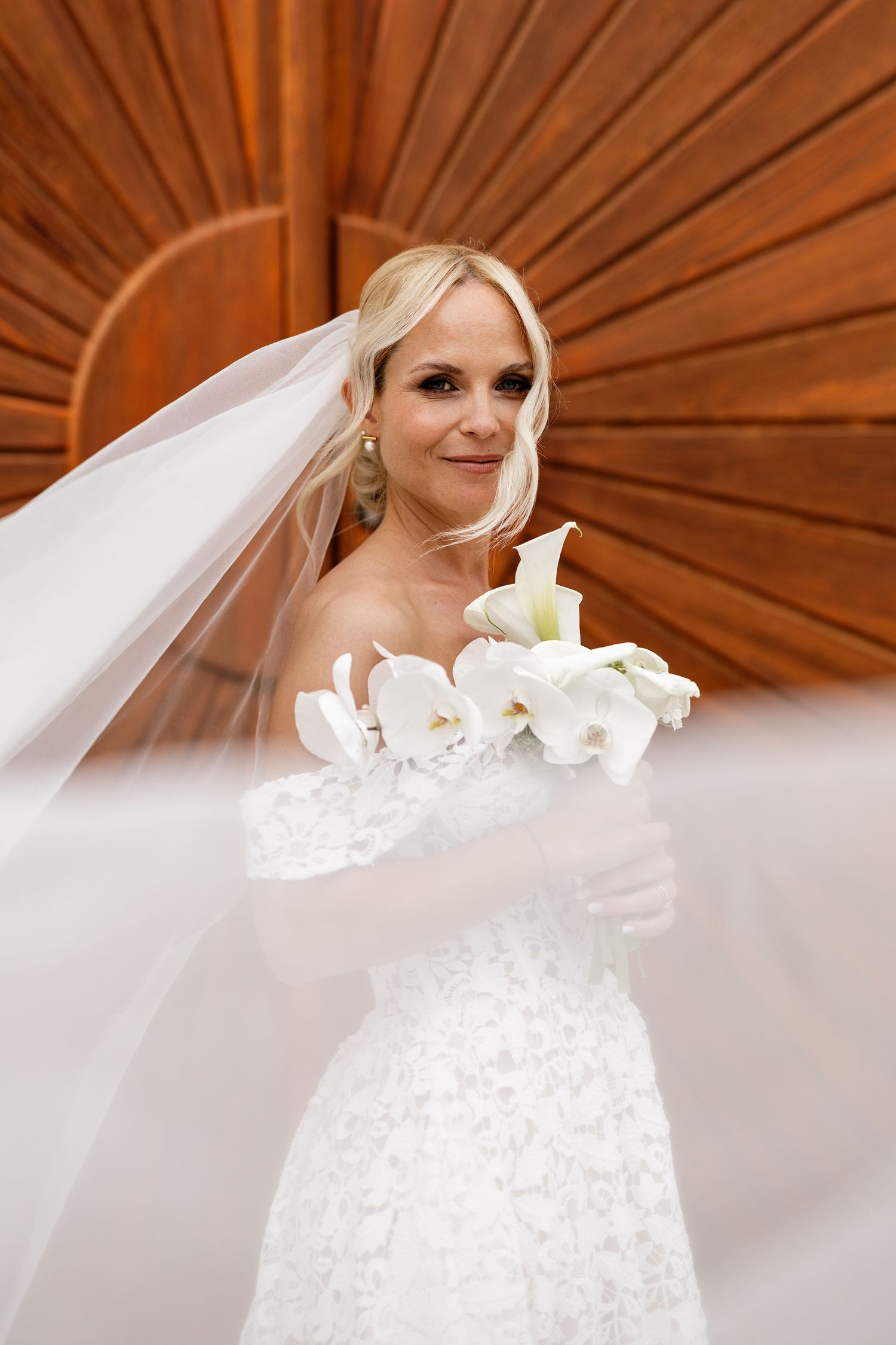 Beautiful blonde bride in lace wedding gown holding white calla lily bouquet with flowing veil against wood backdrop.