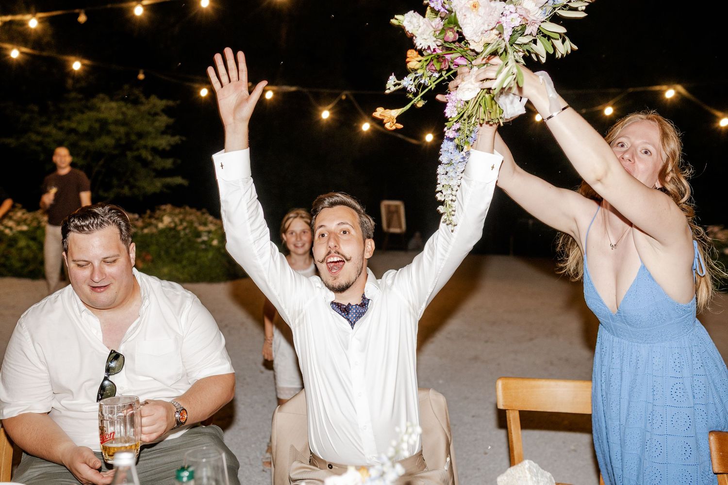 Man triumphantly raises arms at wedding reception under string lights while woman holds bouquet nearby.