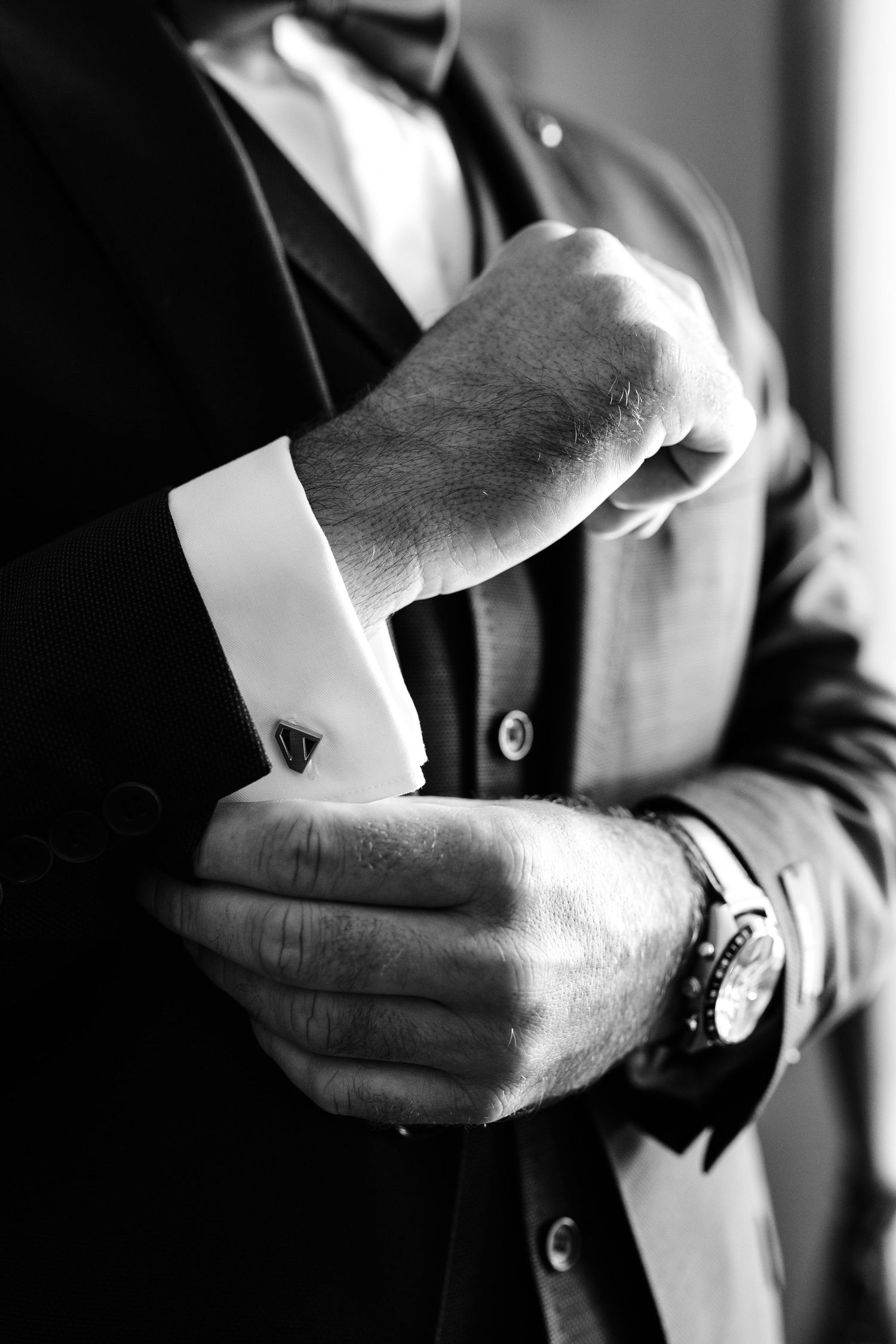 A groom in a black suit adjusting his cufflinks, captured in elegant black and white photography.