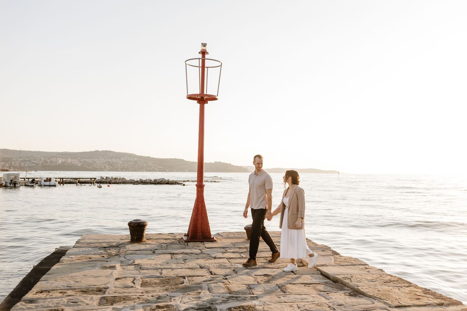 Couple holding hands on a stone pier by the sea near a red lighthouse at sunset.