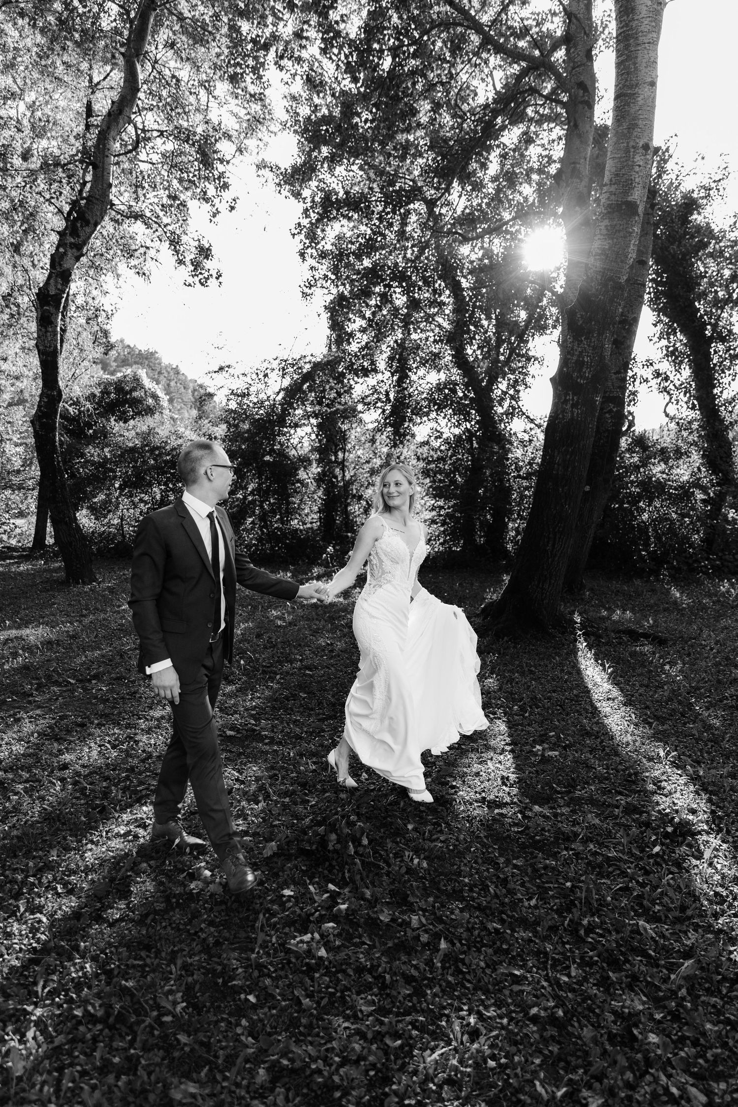 Black and white photo of bride and groom holding hands walking through sunlit forest on wedding day.