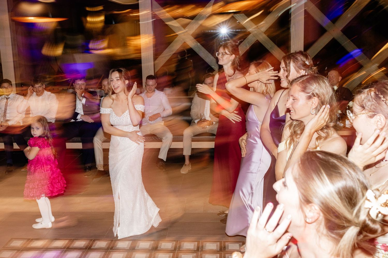 Bride and bridesmaids dancing energetically on wedding reception dance floor with motion blur effect.