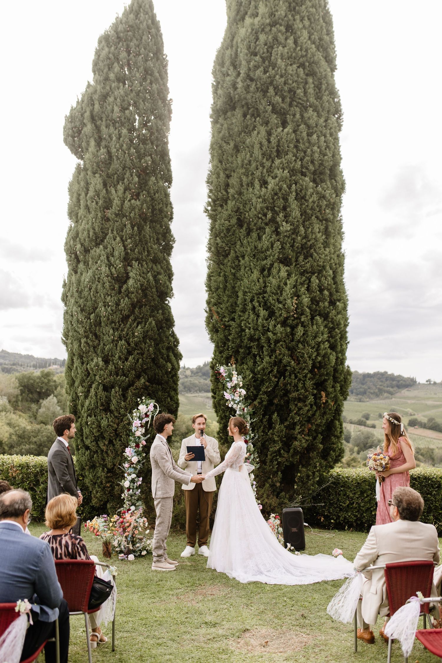 Outdoor wedding ceremony with couple exchanging vows beneath tall cypress trees in a scenic Tuscan countryside setting.