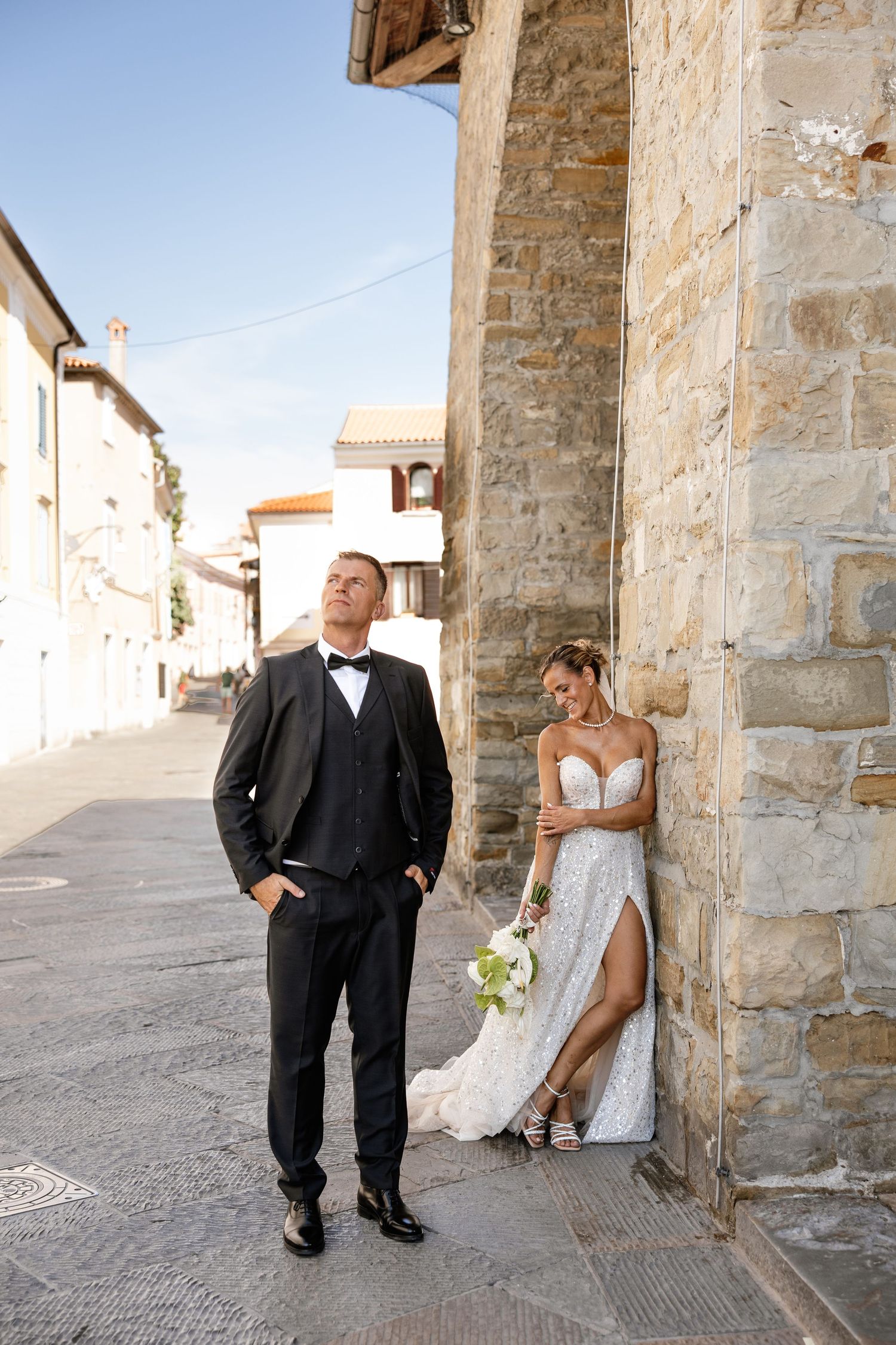 Bride in lace gown and groom in black tuxedo posing on a cobblestone street beside a stone building.