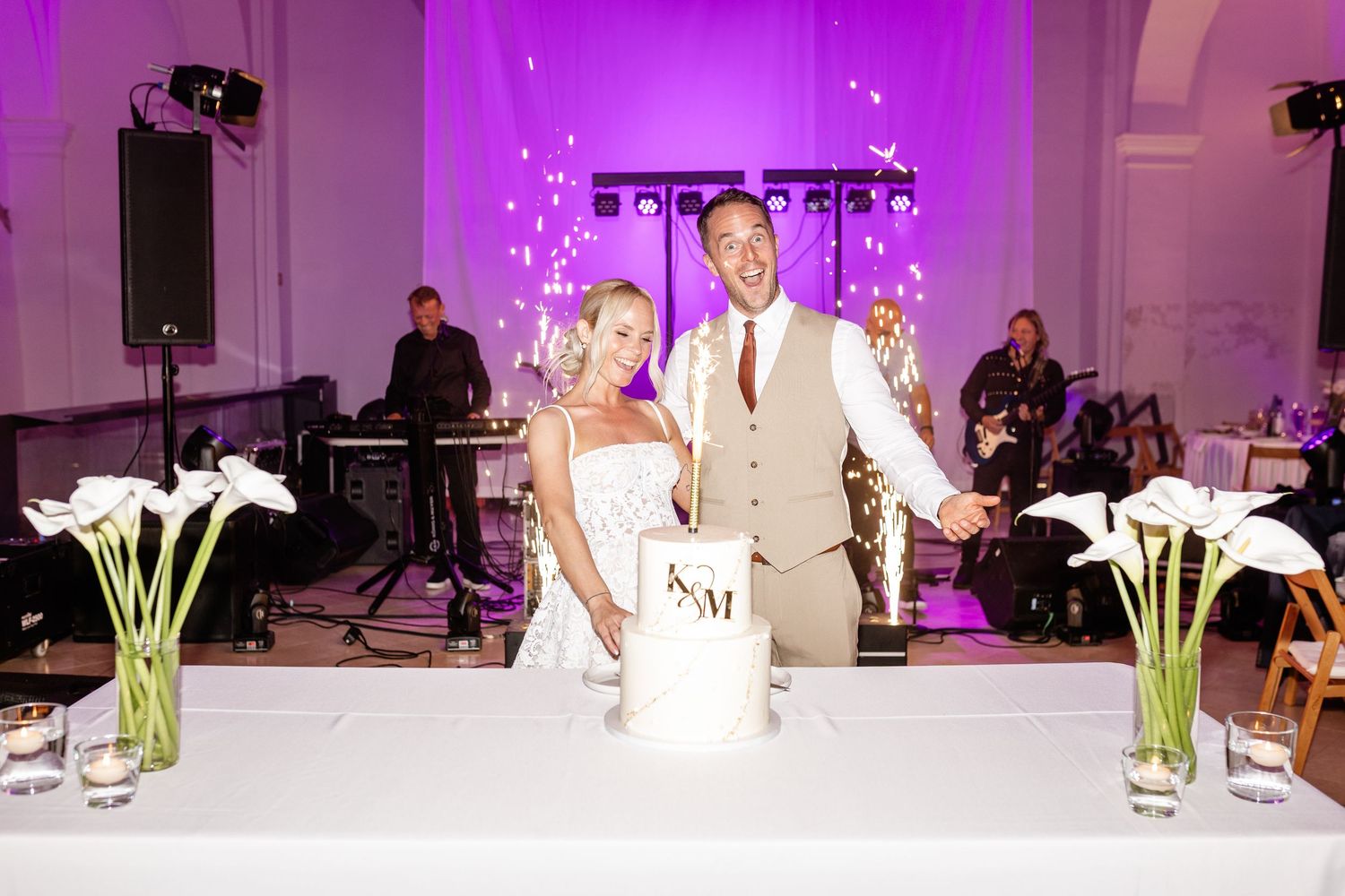 Bride and groom cutting wedding cake at elegant reception with purple lighting and calla lily centerpieces.