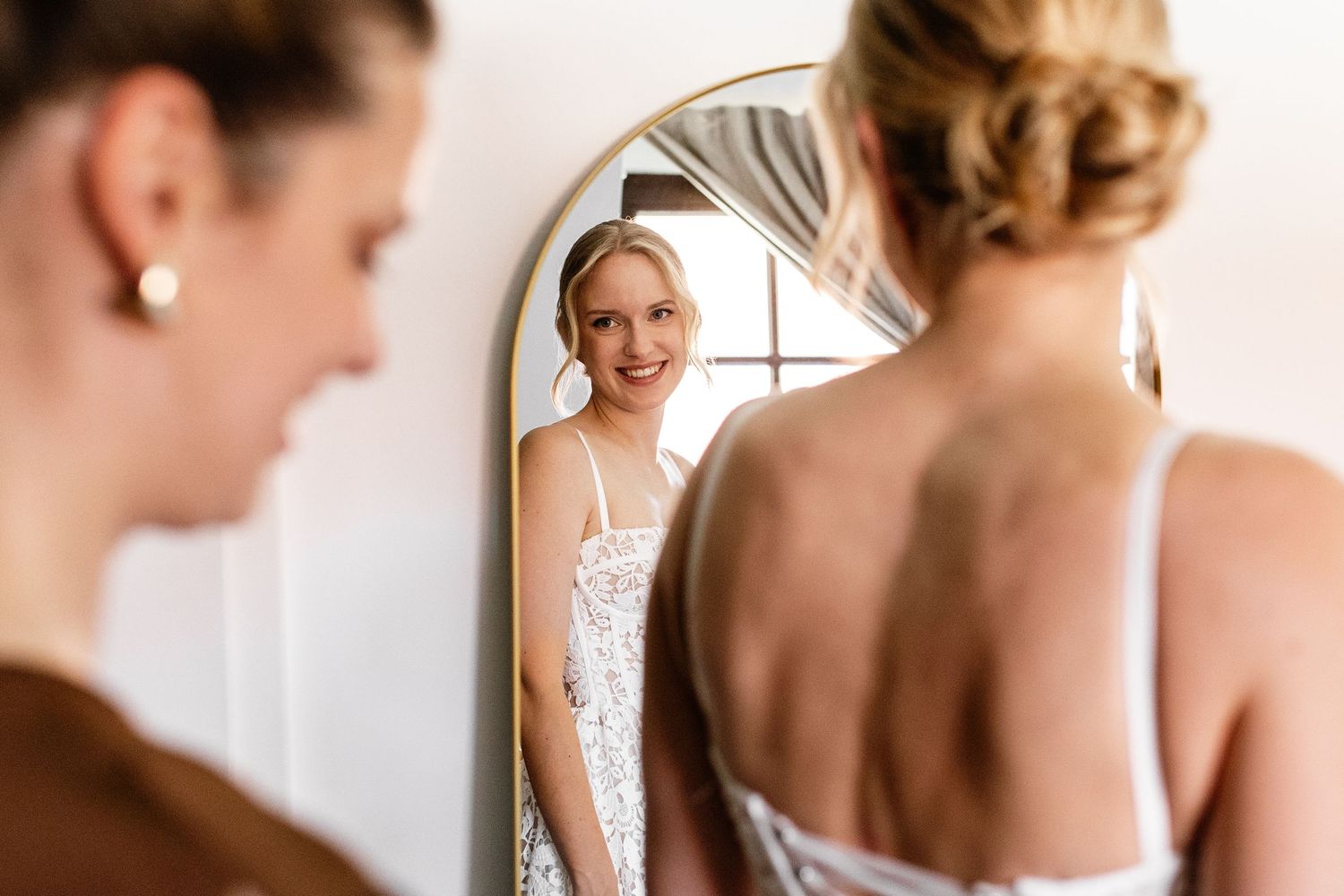 Bride in lace gown admires her reflection in an arched gold mirror while getting ready on her wedding day.