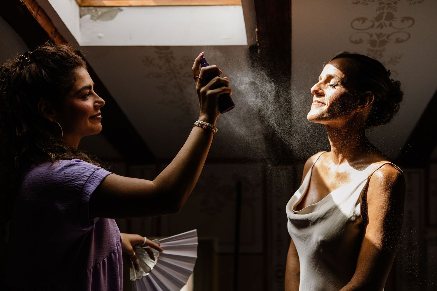 A makeup artist sprays setting spray on a bride in a white dress while holding a fan in a softly lit room.
