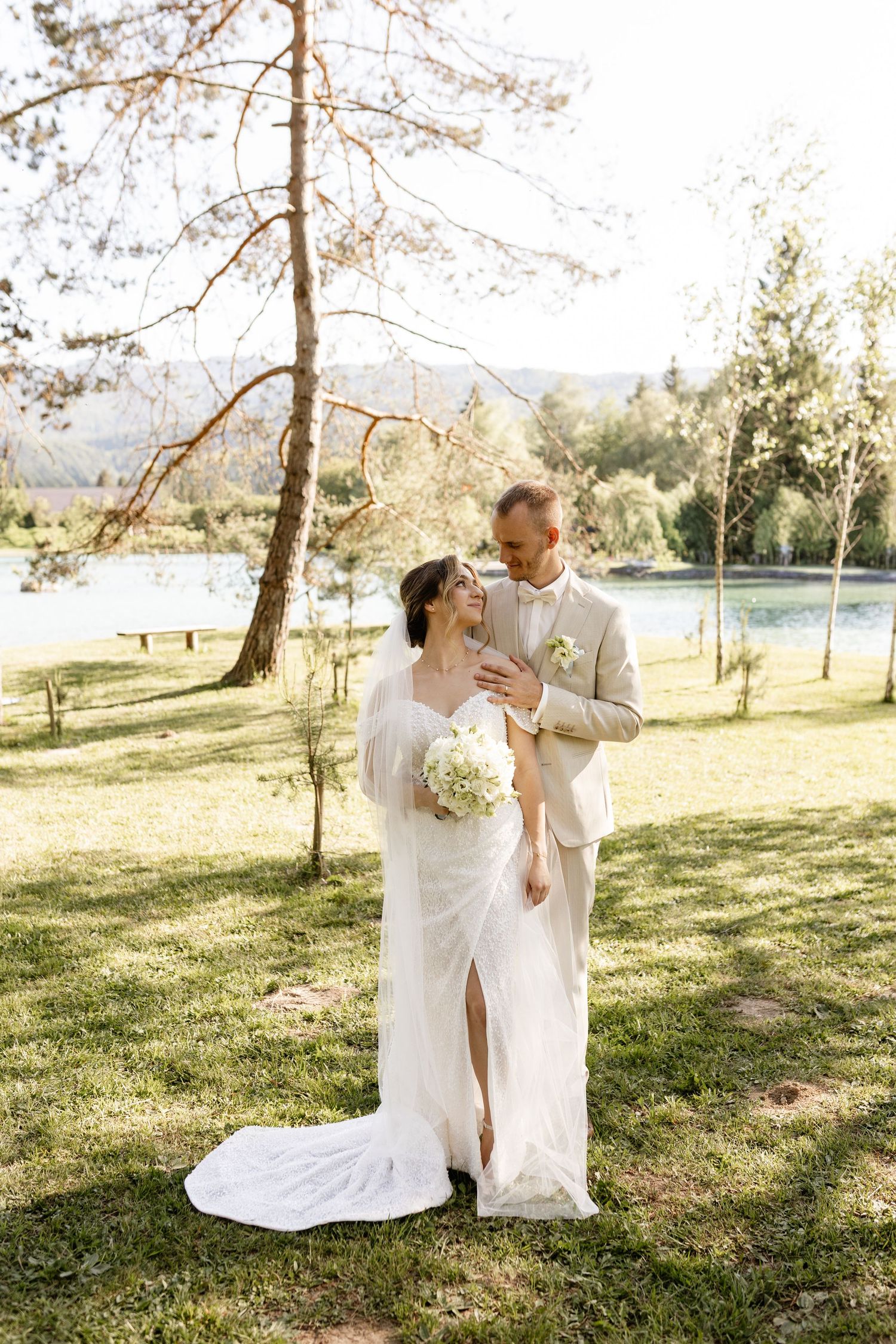 Bride and groom embrace outdoors near a sunlit lake, surrounded by trees, as bride holds a white floral bouquet.