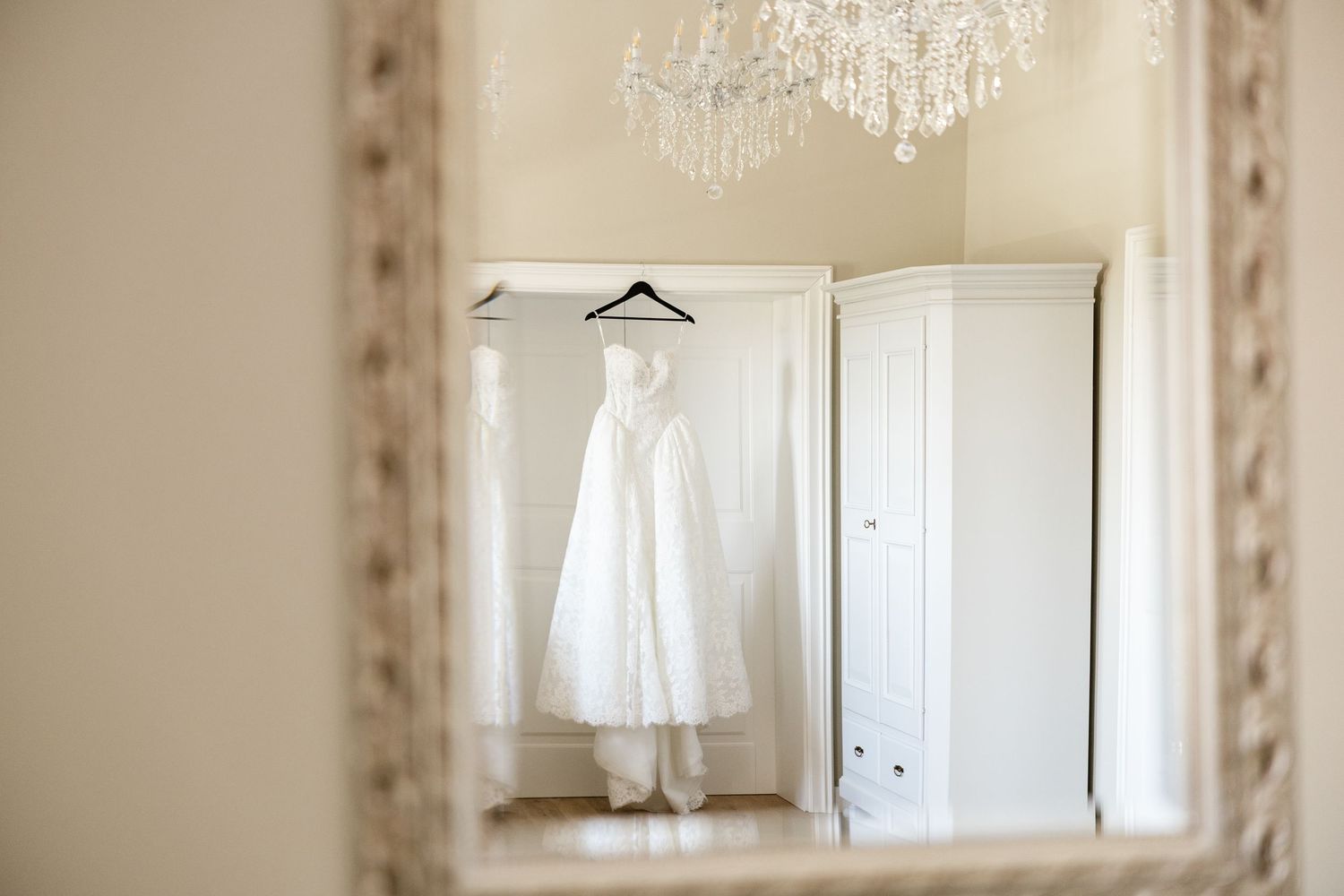 White wedding dress hanging on a hanger reflected in an ornate mirror with a crystal chandelier above.
