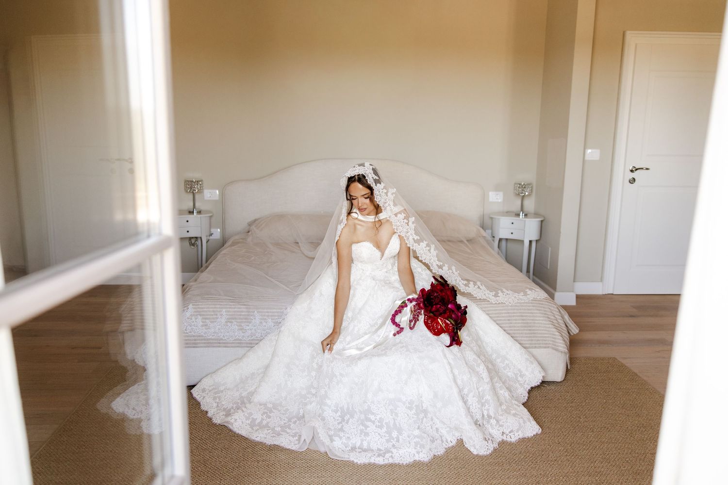 Bride in elegant lace wedding gown with veil sits on bed holding red bouquet in bright bedroom.