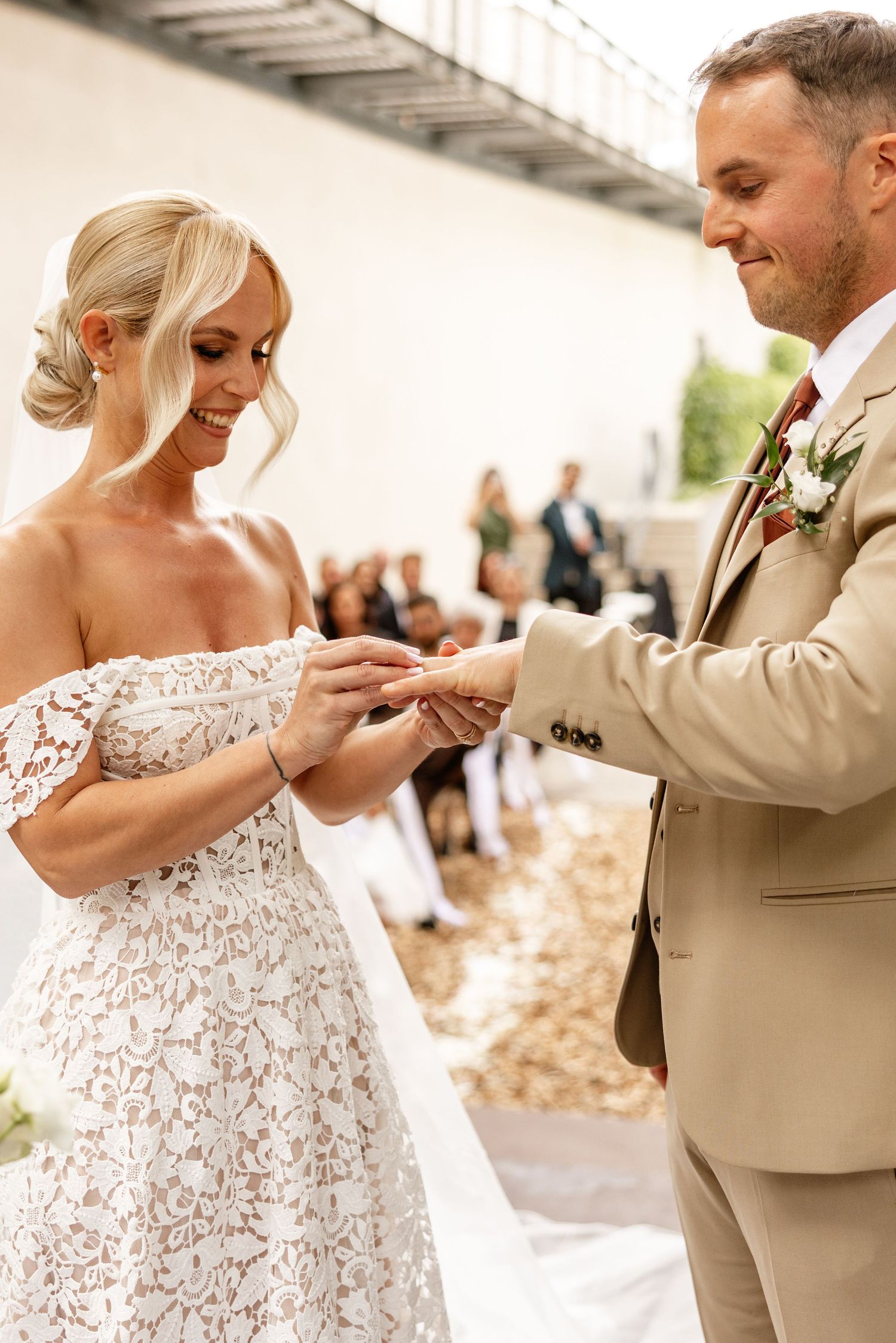 Smiling bride in lace off-shoulder dress receives wedding ring from groom in tan suit during outdoor ceremony.