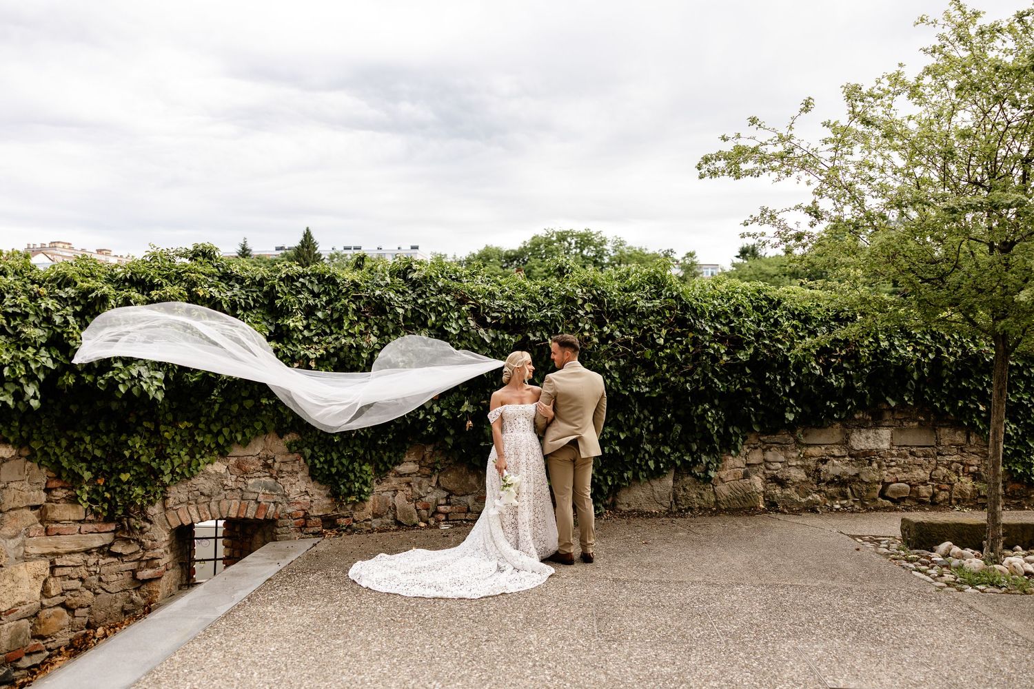 Bride and groom embrace as wedding veil dramatically billows in wind against ivy wall.