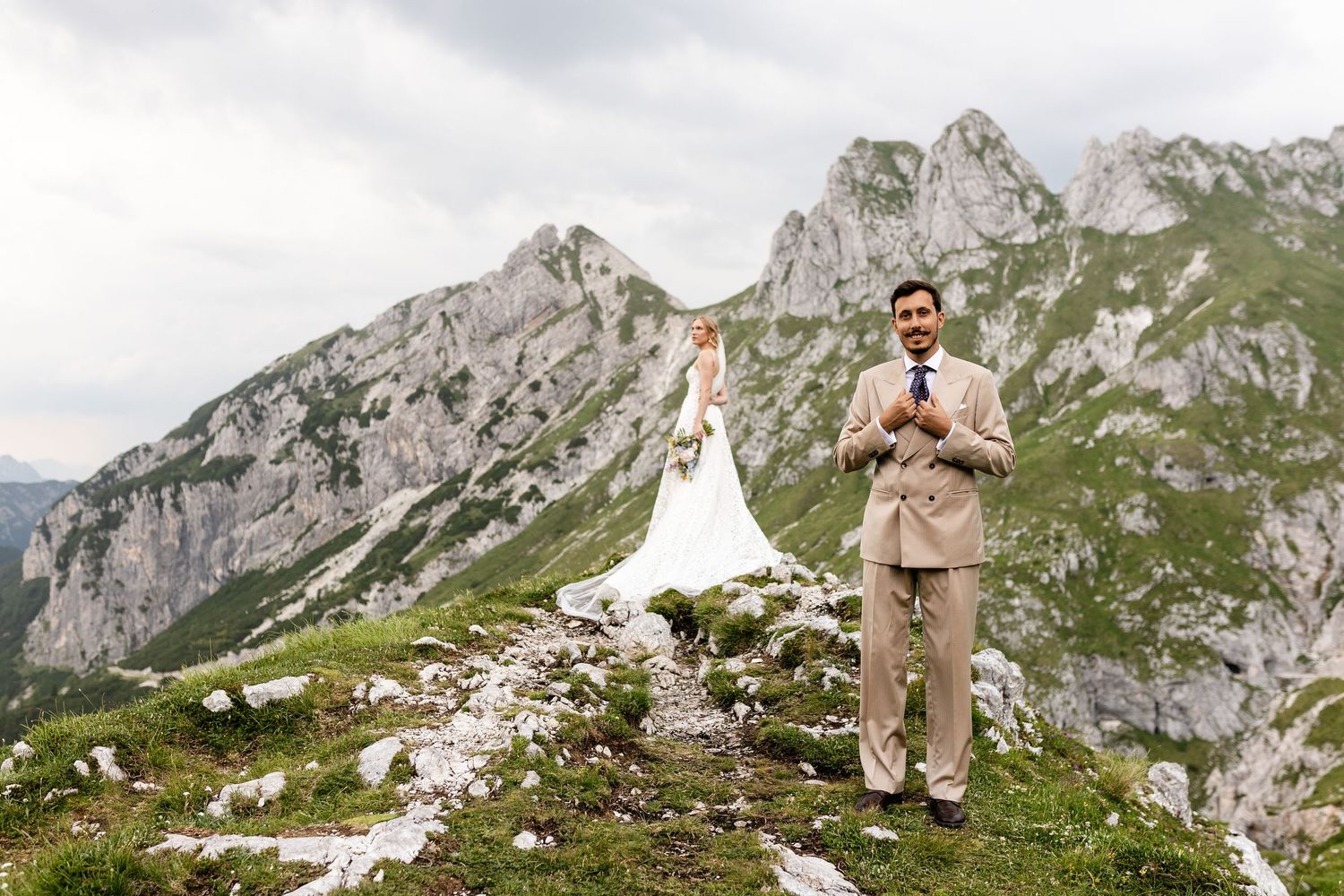 Groom in tan suit stands on rocky Alpine mountain ridge with bride in lace gown posing behind him.