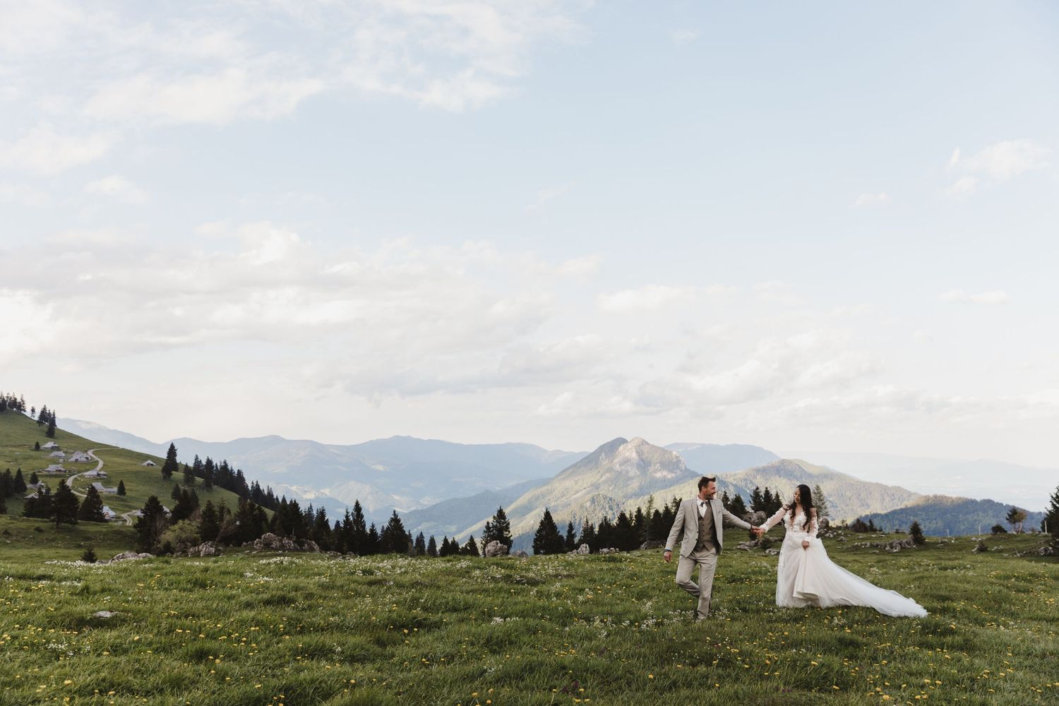 Wedding couple holding hands walking across green alpine meadow with stunning mountain backdrop.