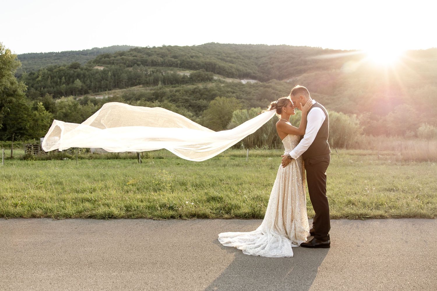 Bride and groom kissing at sunset on a road, brides veil flowing dramatically in the wind with hills behind.