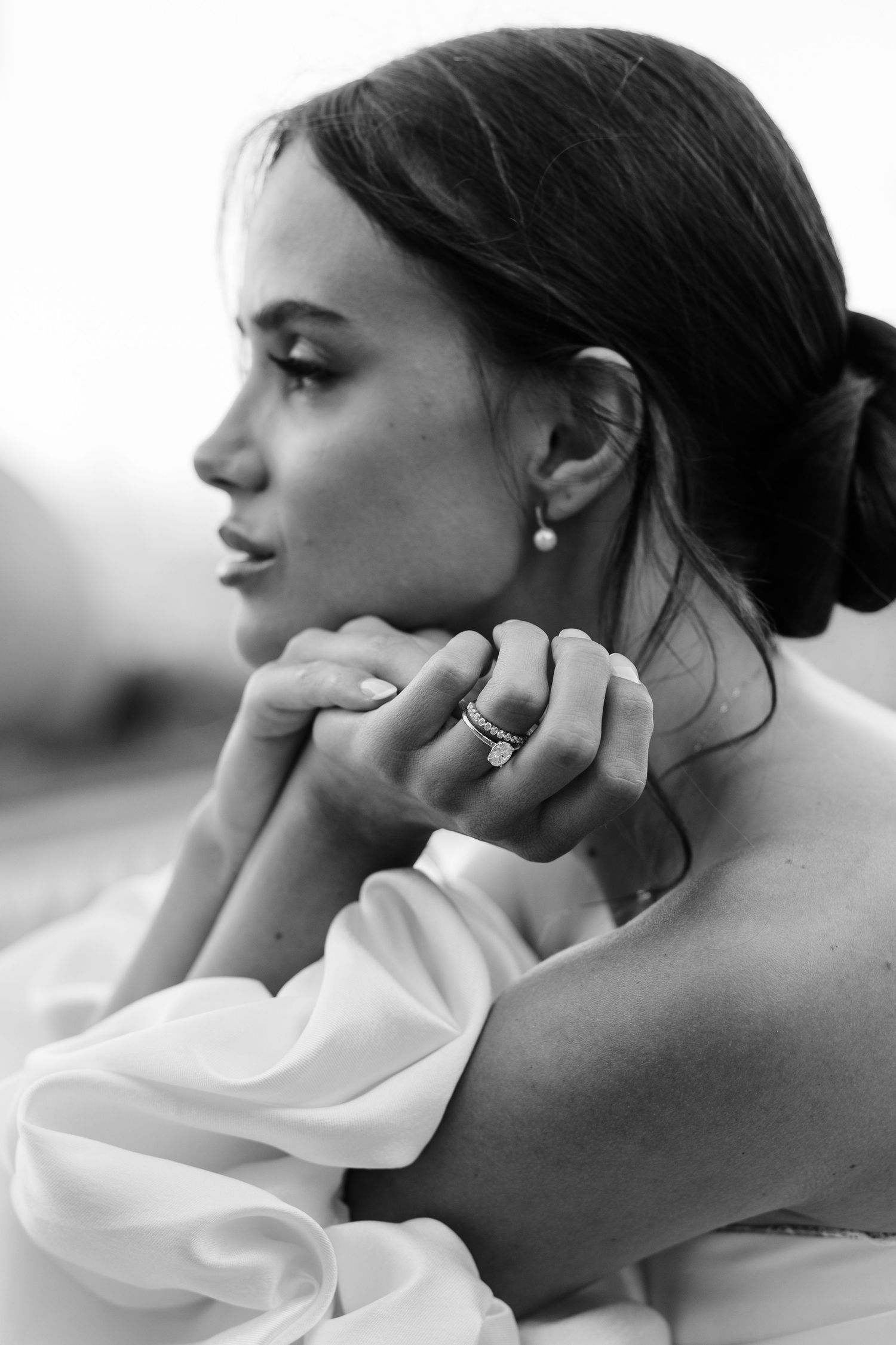 Elegant bride in off-shoulder gown poses thoughtfully, showcasing pearl earring and stacked rings.