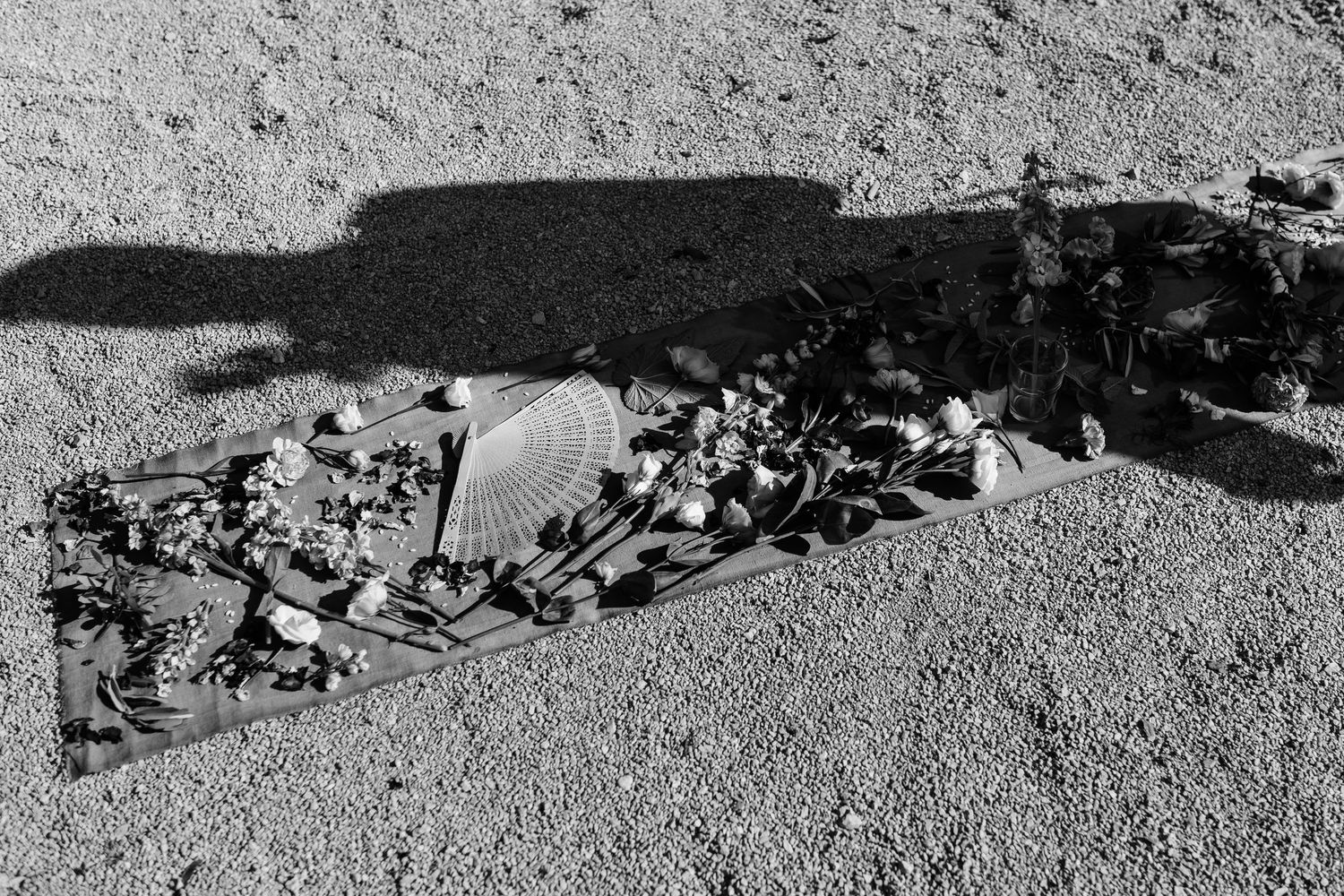 Black and white photo of flowers and decorative items arranged on a cloth on a gravel surface.