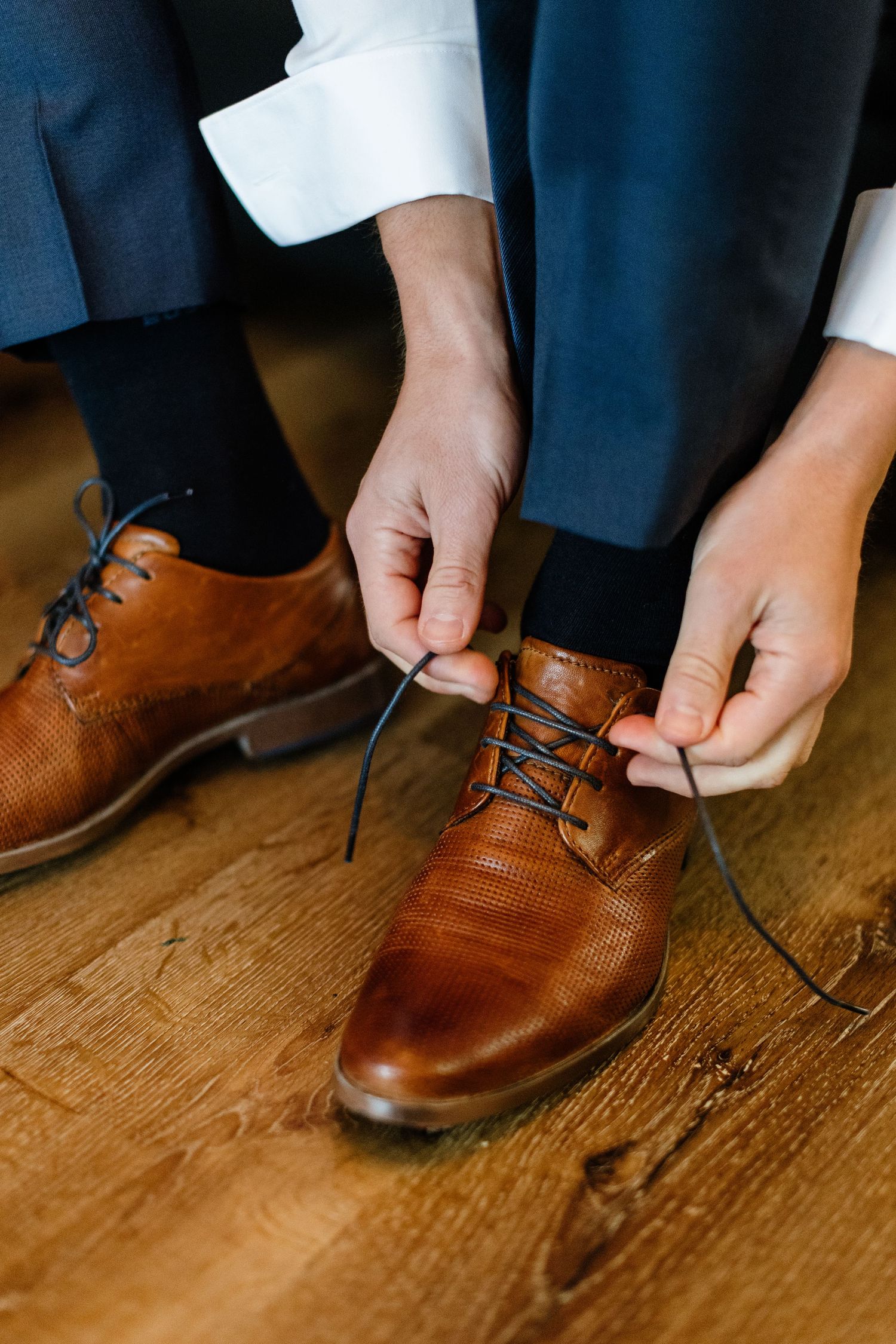Man tying blue laces on brown leather oxford dress shoes while wearing gray trousers and dark socks.