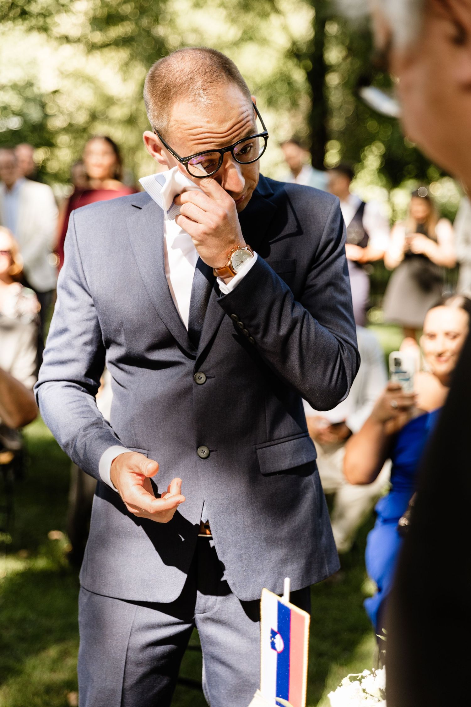 Emotional man in a suit wiping tears at an outdoor wedding ceremony.