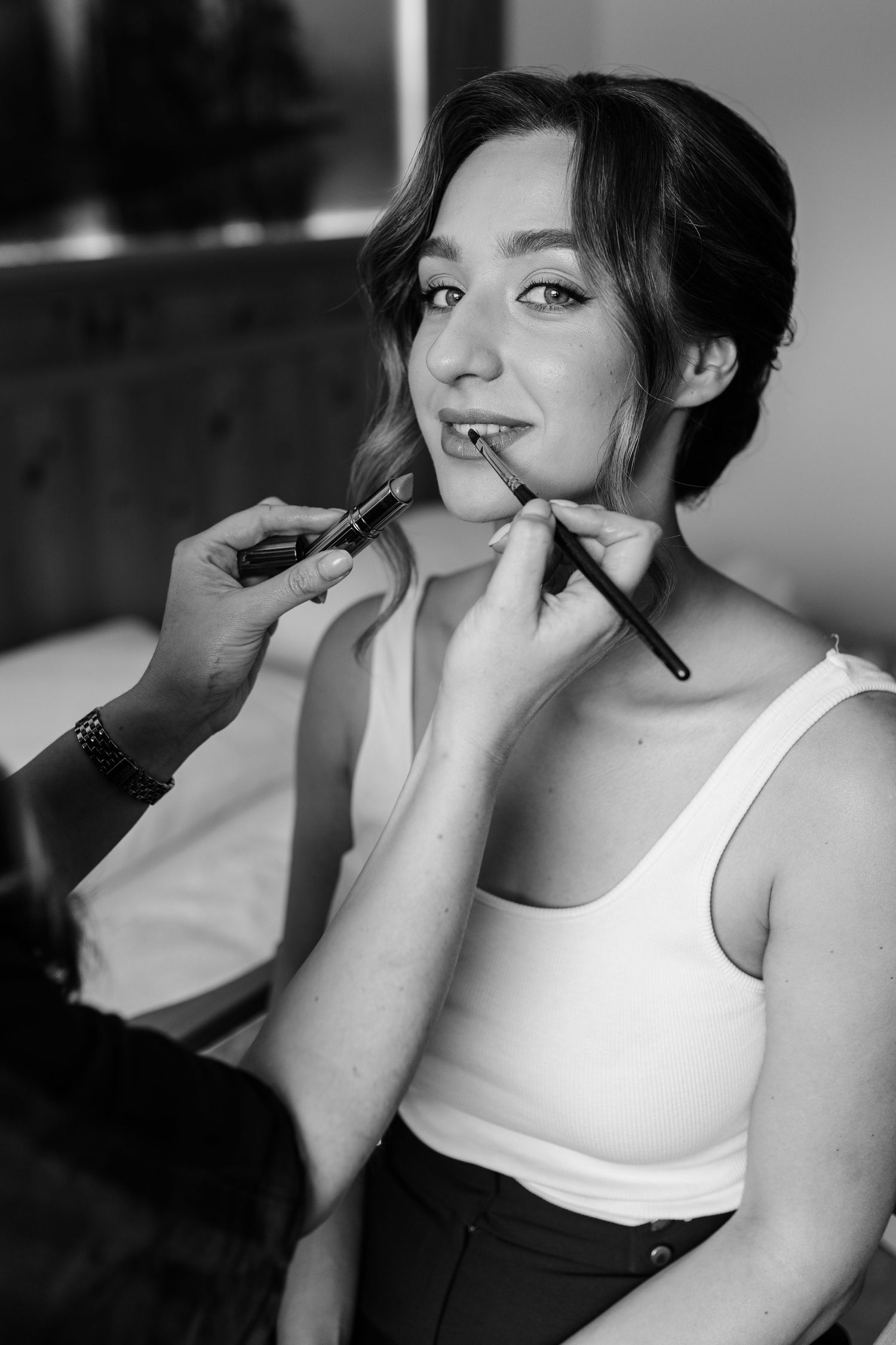 Black and white photo of a bride getting makeup applied with two brushes on her lips before her wedding.