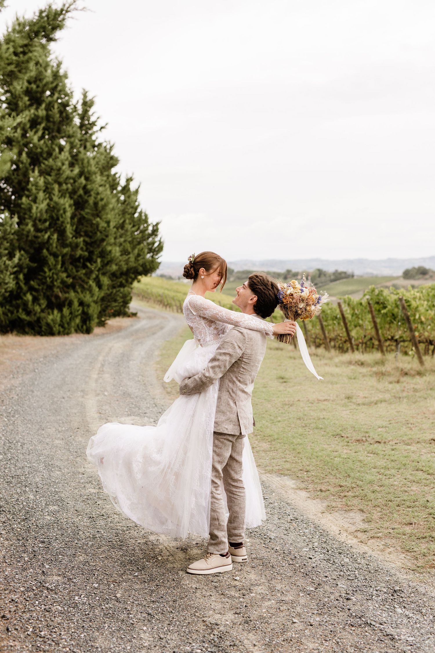 Groom lifts bride in white lace dress on a scenic vineyard path surrounded by cypress trees.
