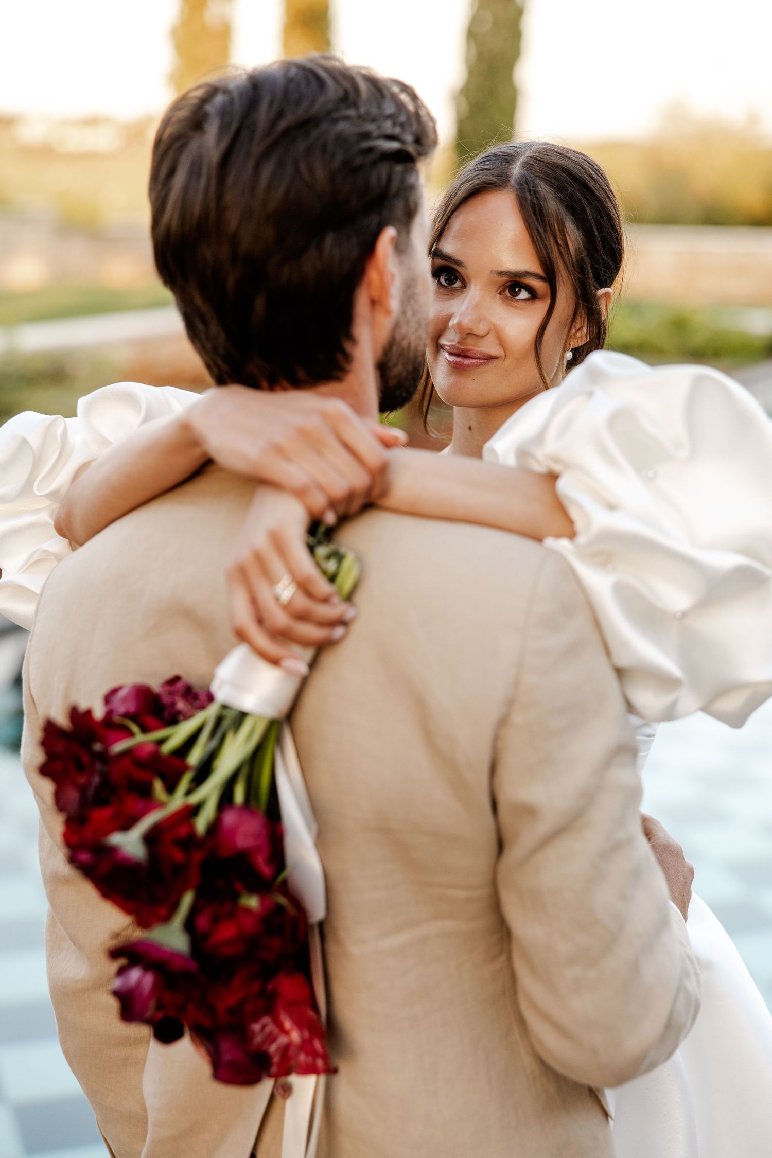 Bride in white puffed sleeve gown embraces groom holding red tulip bouquet during golden hour outdoor wedding.