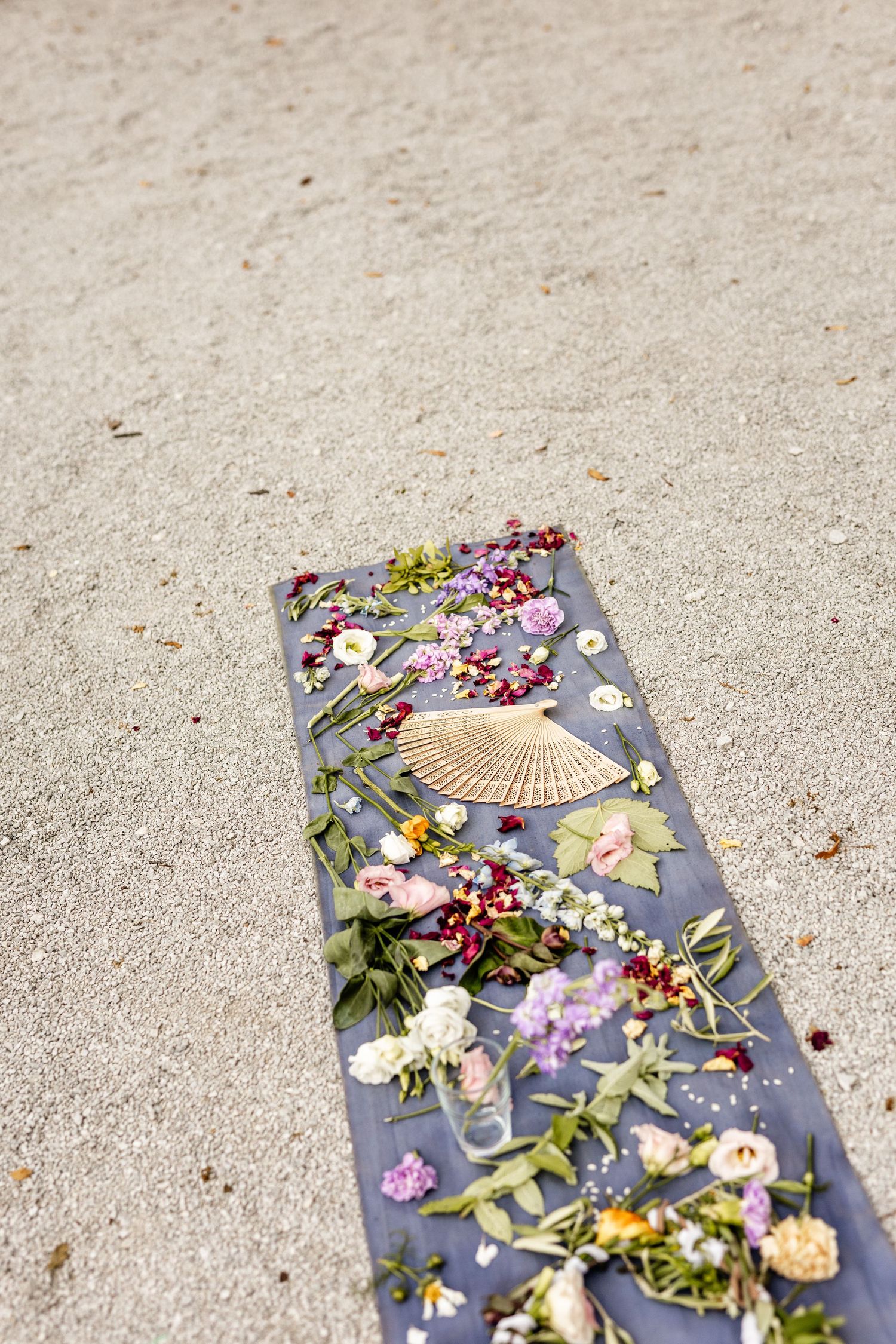 Colorful wedding ceremony flower petals, greenery, and a fan arranged on gray fabric runner on sandy ground.