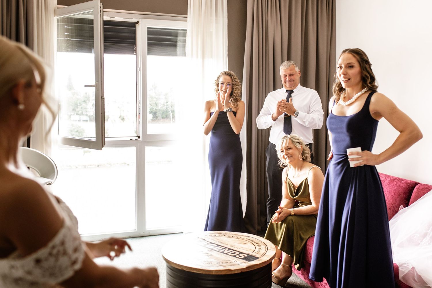 Bridesmaids in navy dresses react with joy as bride gets ready in bright hotel room with guests watching.