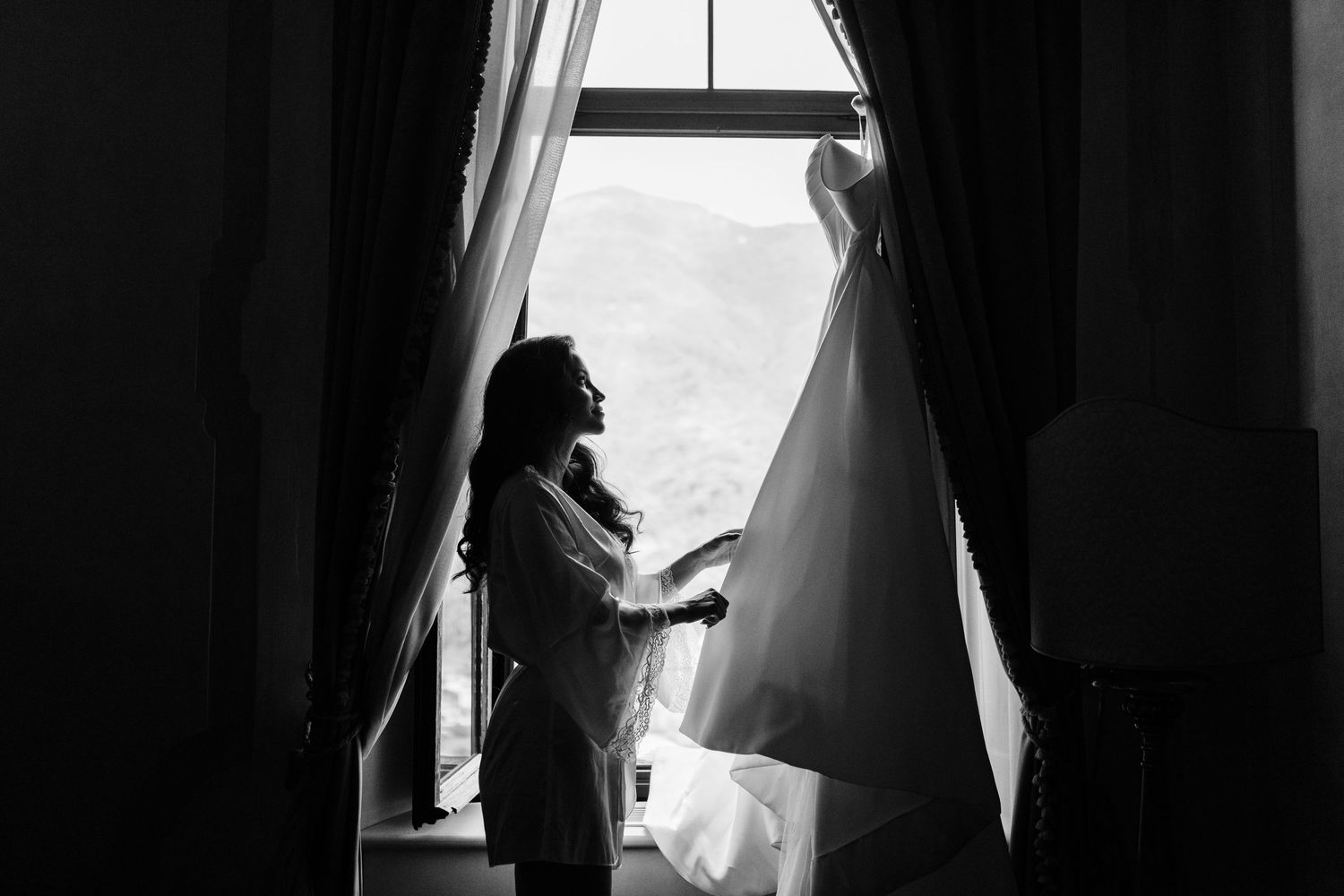 Black and white silhouette of bride holding wedding dress near window with mountain view.