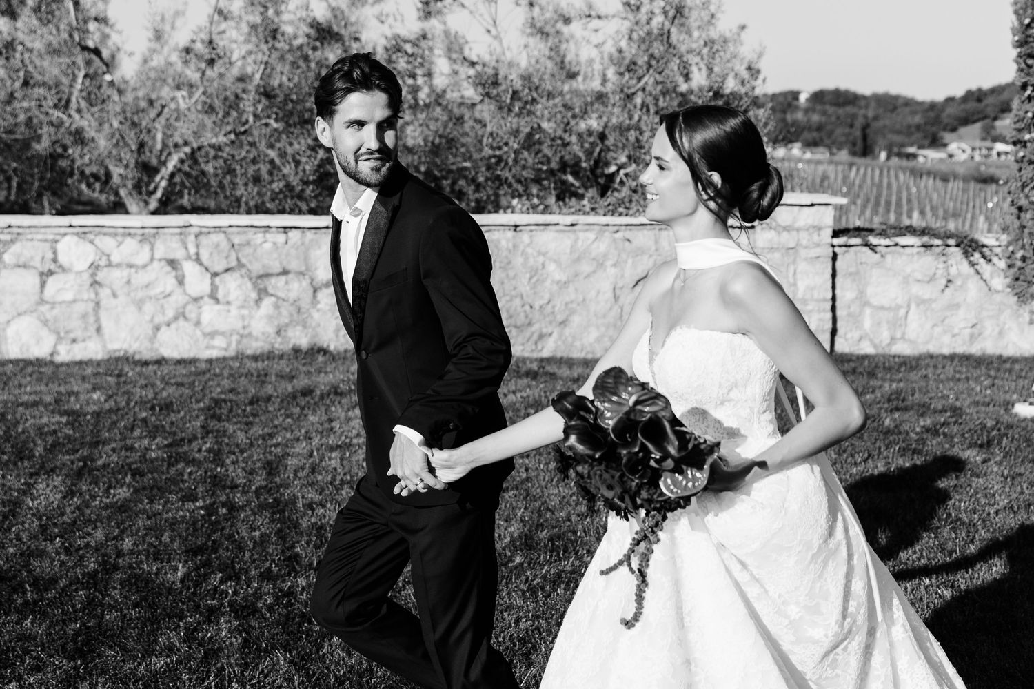 Black and white photo of bride and groom walking hand in hand along a stone garden wall.