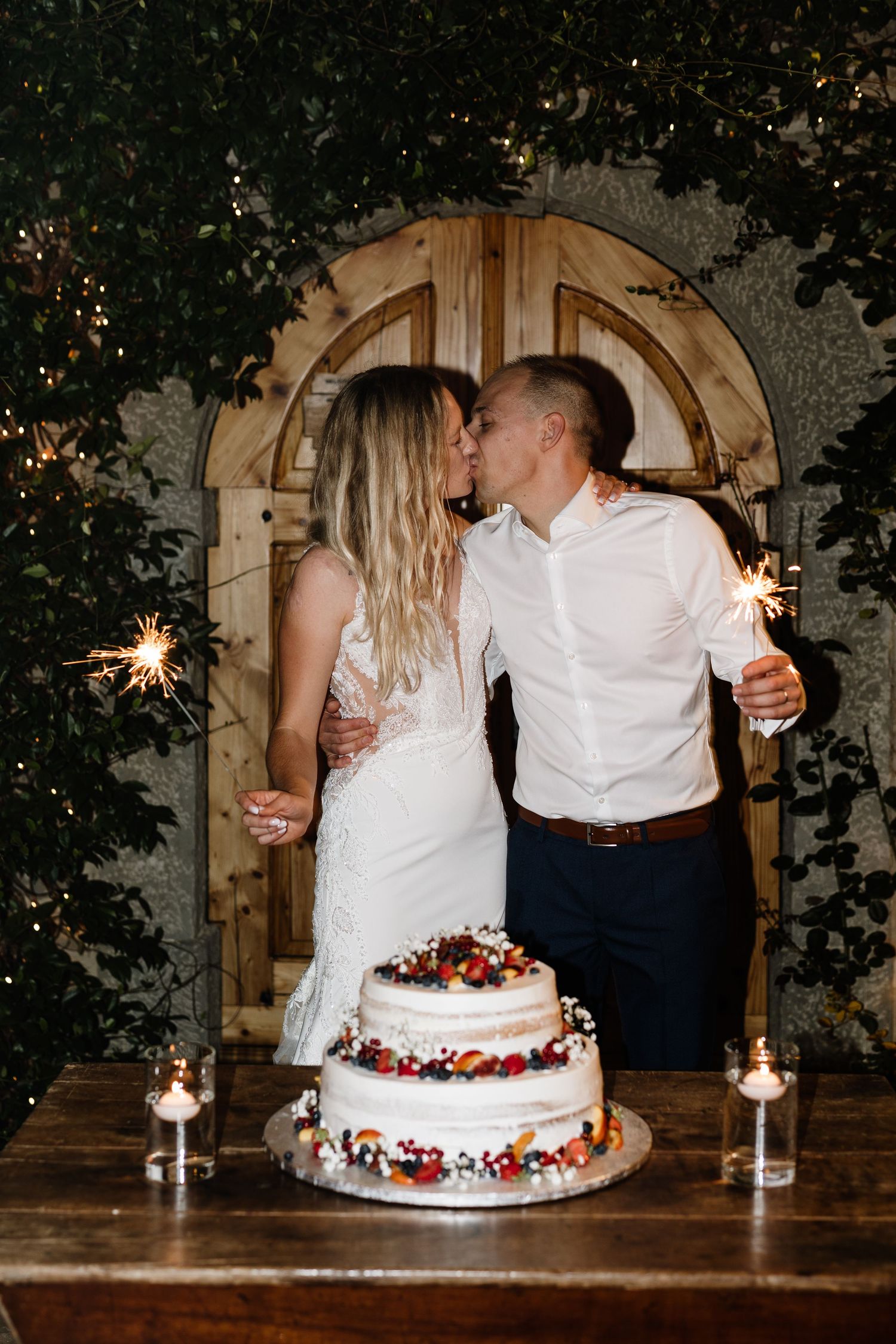 Bride and groom kissing beside a decorated two-tier wedding cake holding sparklers.