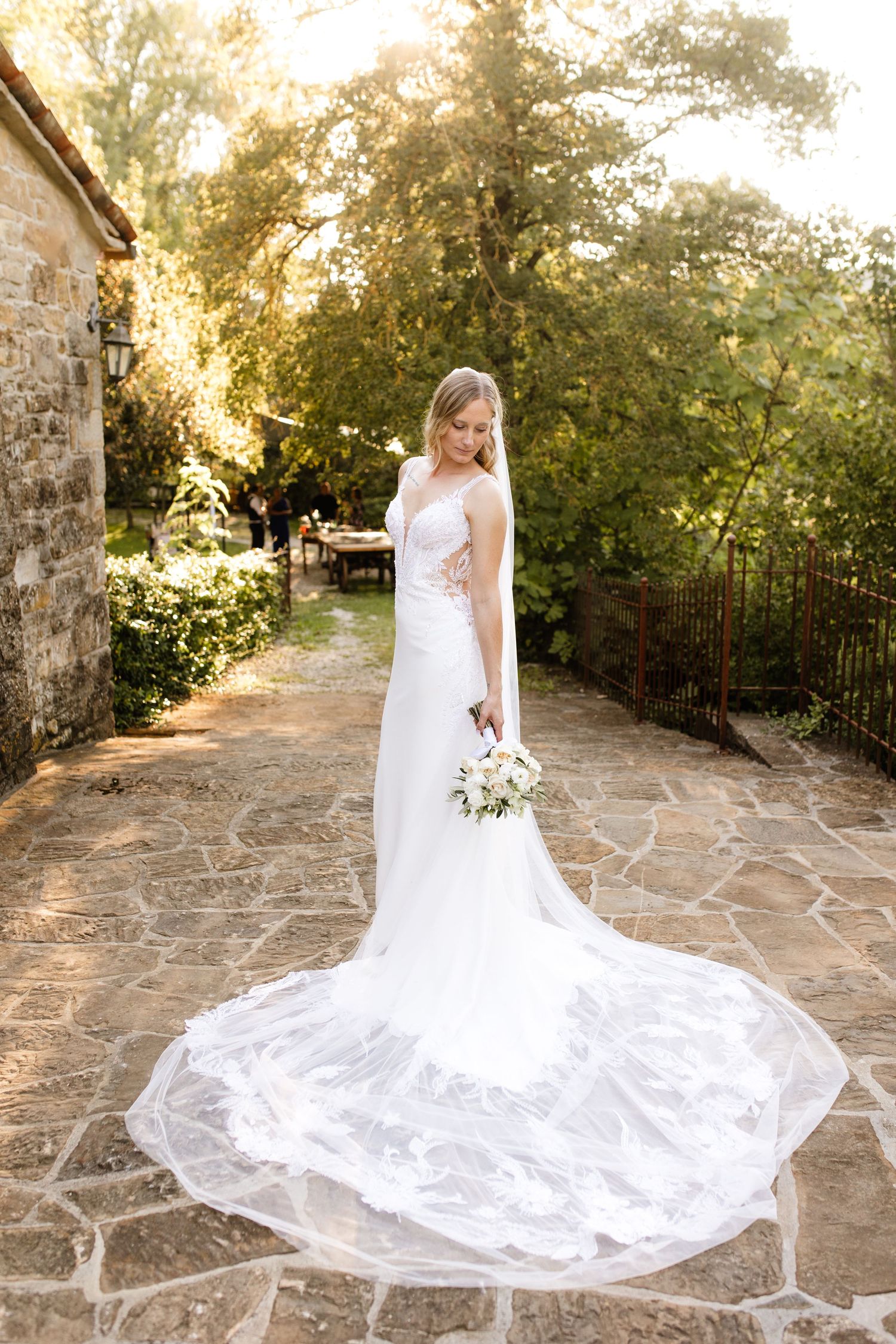 Elegant bride in white gown with lace train holds bouquet on stone patio surrounded by lush greenery.