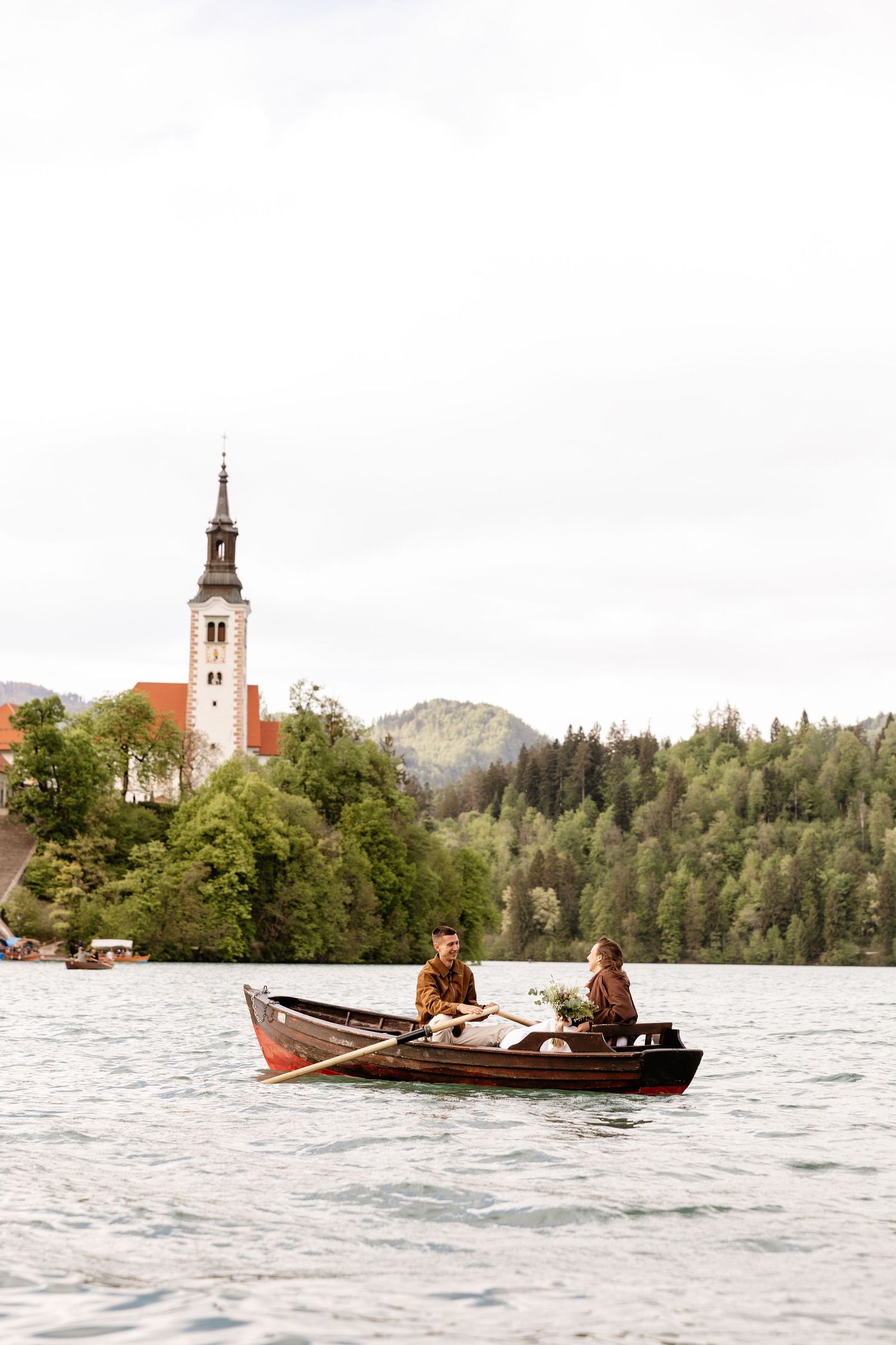 Couple rowing a wooden boat on Lake Bled with a church tower and lush green forest in the background.