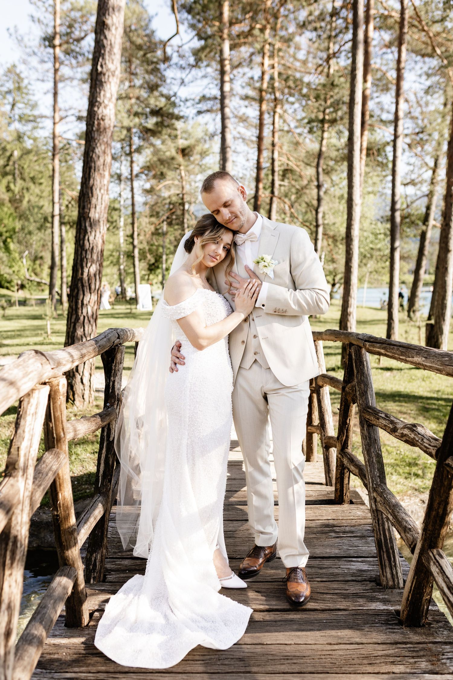 Bride and groom embracing on a wooden bridge surrounded by tall pine trees on a sunny wedding day.