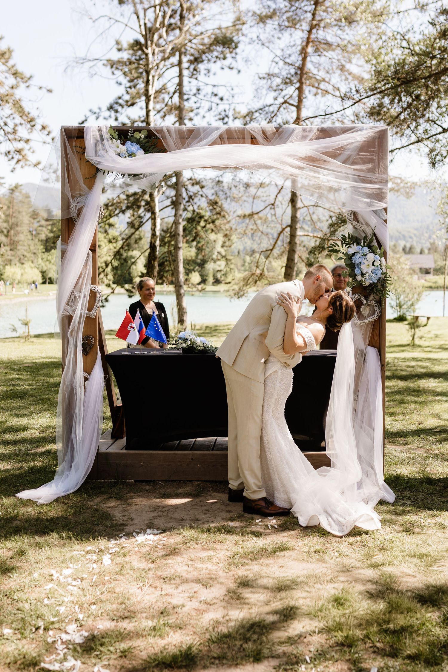 Bride and groom share first kiss under decorated outdoor wedding arch surrounded by trees and nature.