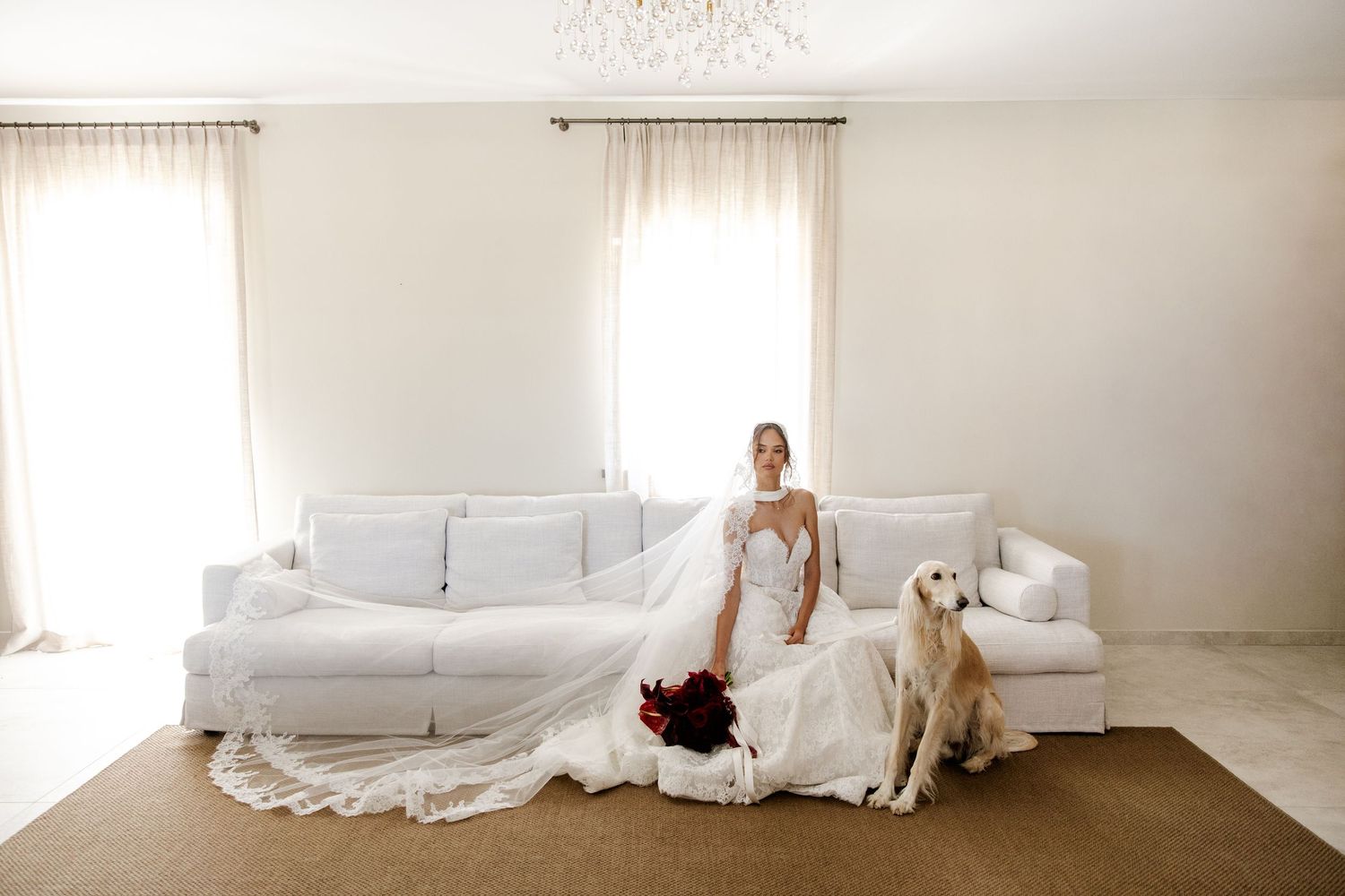 Bride in white gown sits with golden retriever beside white sofa, holding red bouquet in bright elegant room.